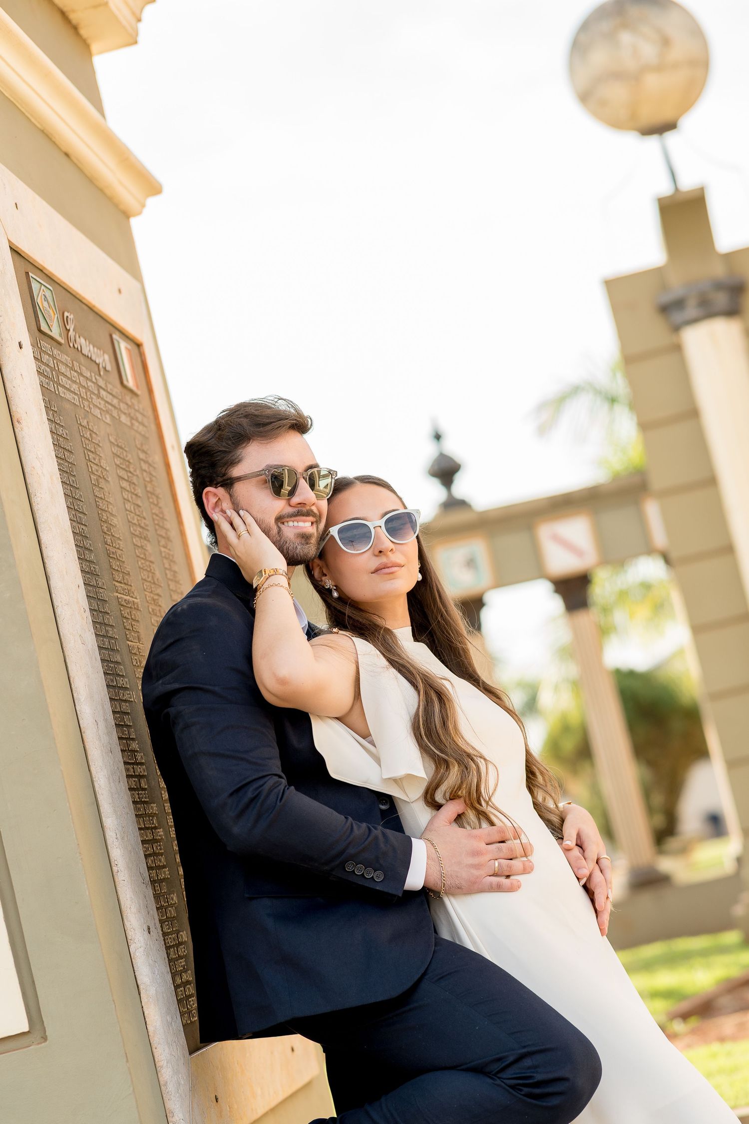 Couple in sunglasses posing outdoors, man in navy suit embracing woman in white dress near decorative pillar.