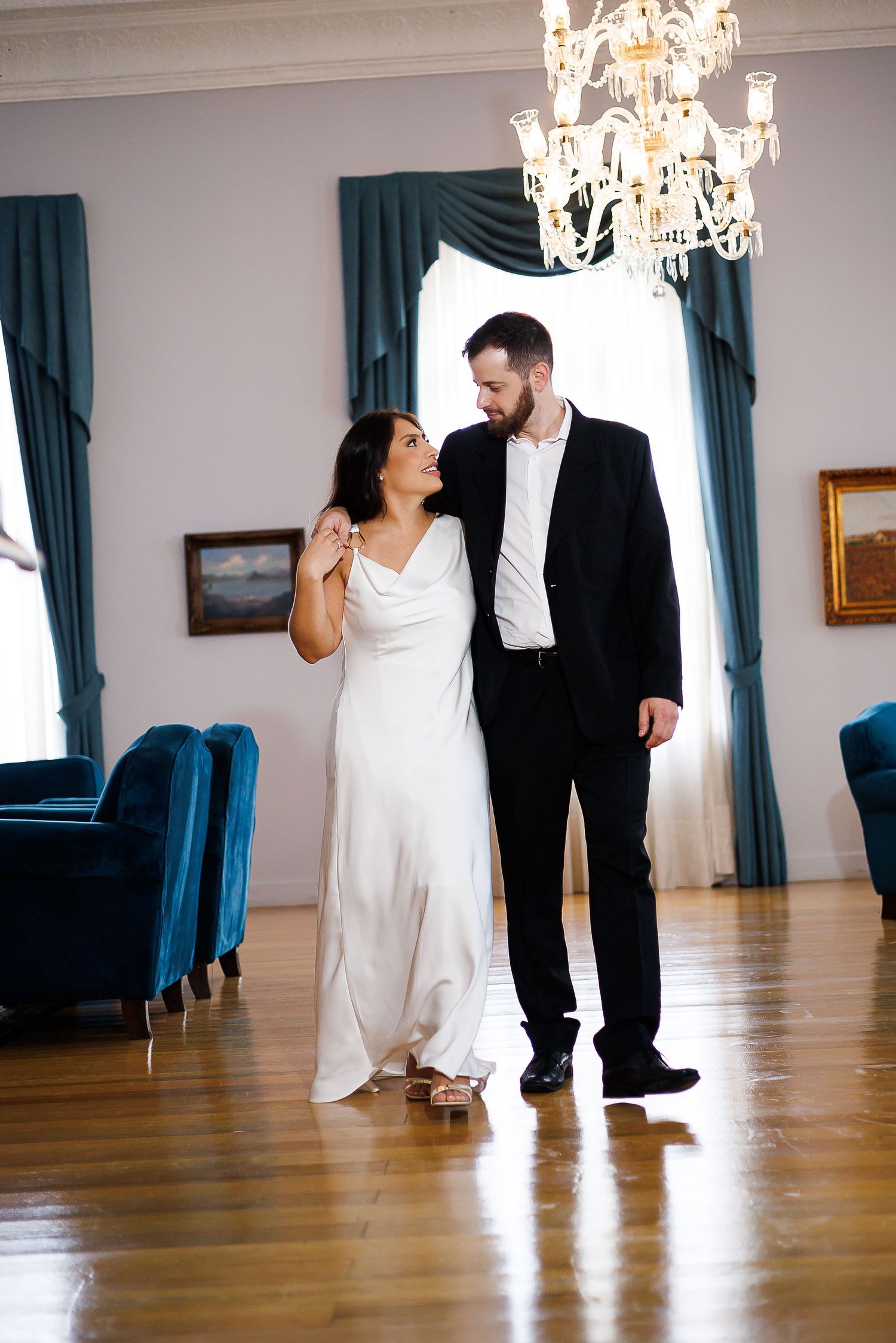 Bride in white gown and groom in black suit share a romantic moment in an elegant ballroom with chandelier.