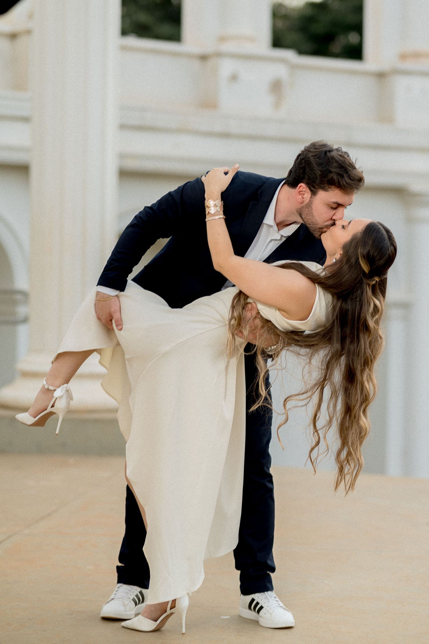 A groom dips his bride for a romantic kiss outdoors, she wears a white dress and heels, he wears a navy suit.