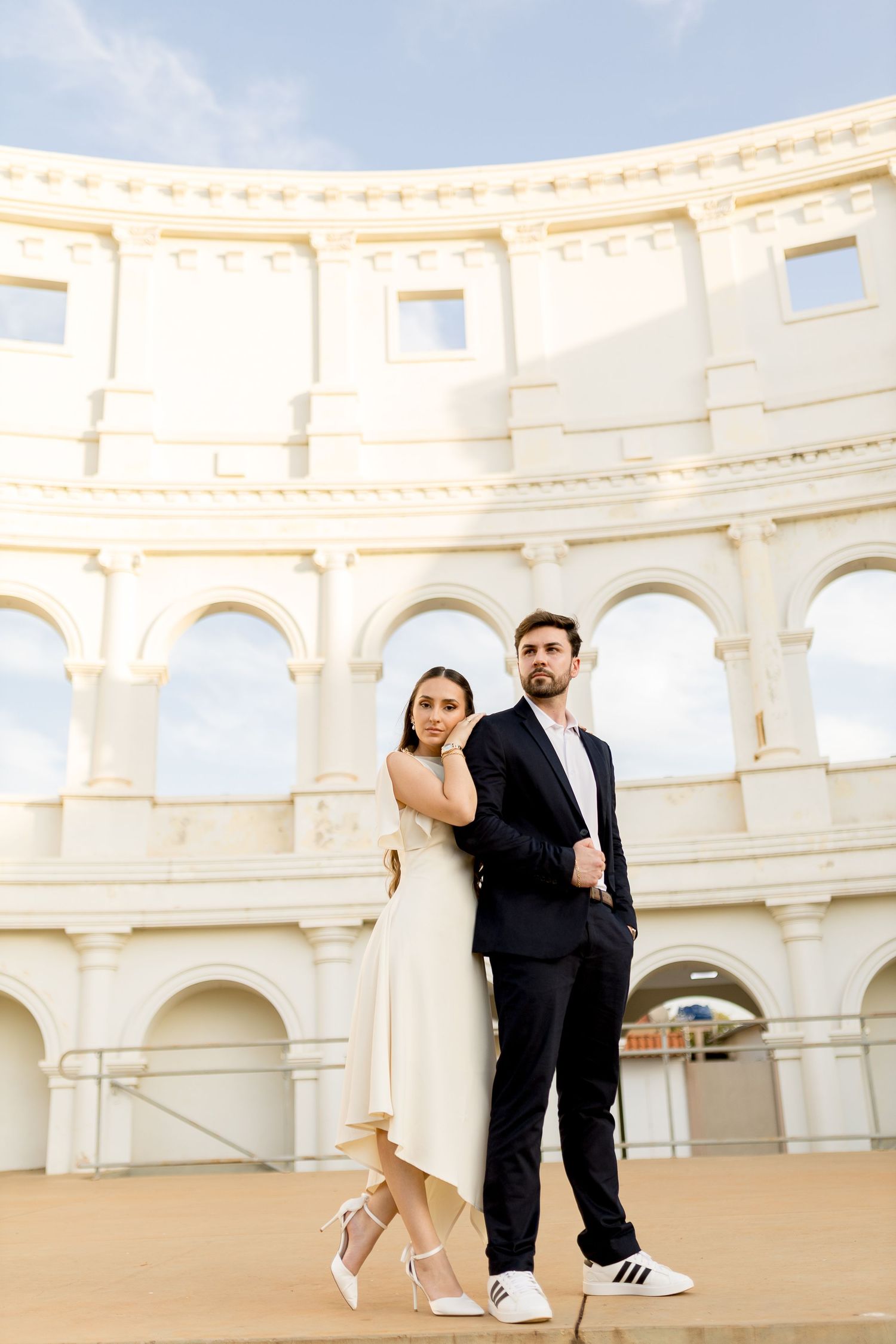 Elegant couple posing in front of a white Roman amphitheater, woman in cream dress, man in dark suit.