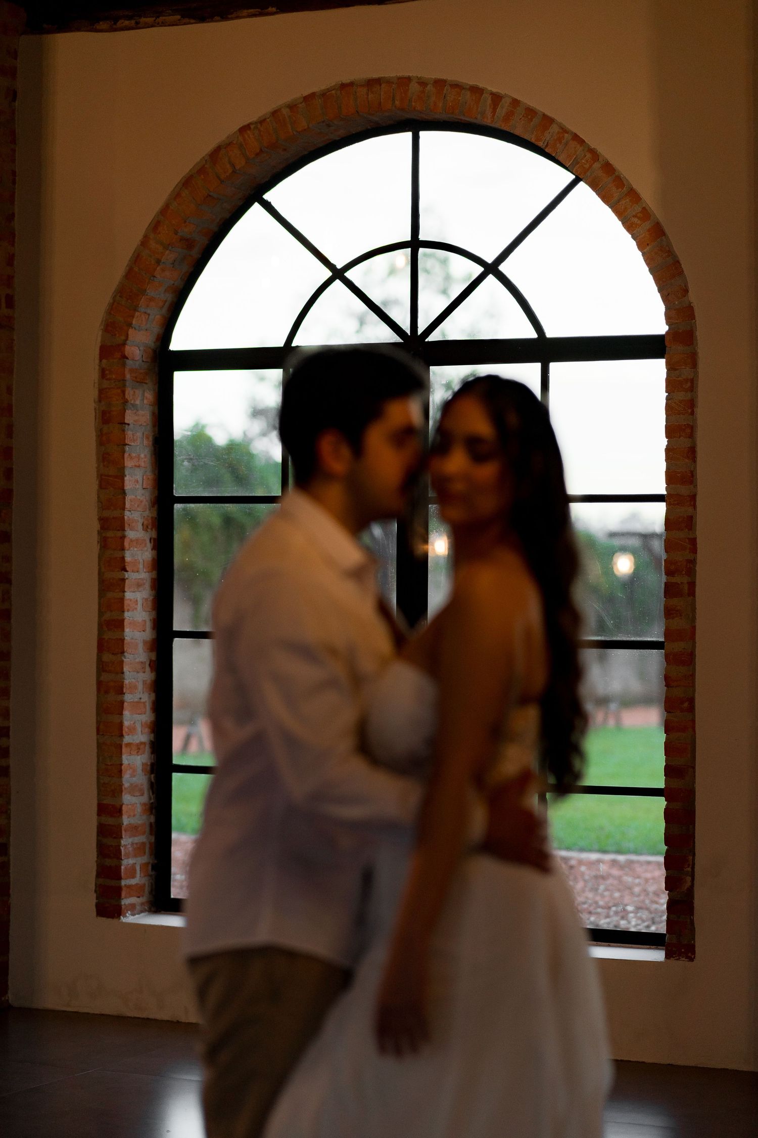 Couple embracing in front of arched brick window with garden view, romantic indoor portrait in soft focus.