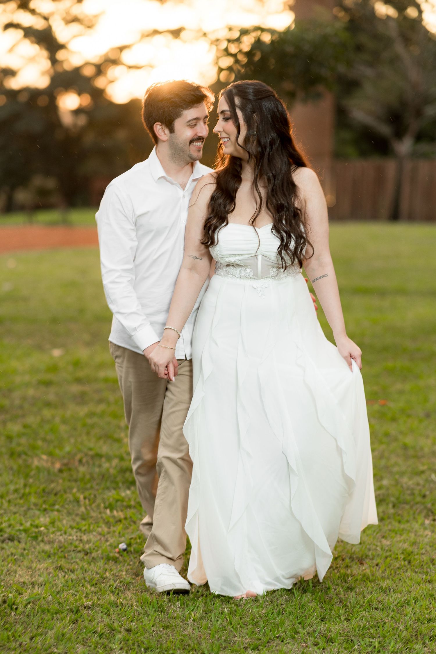 A couple in wedding attire walks hand-in-hand on grass at sunset, smiling at each other.