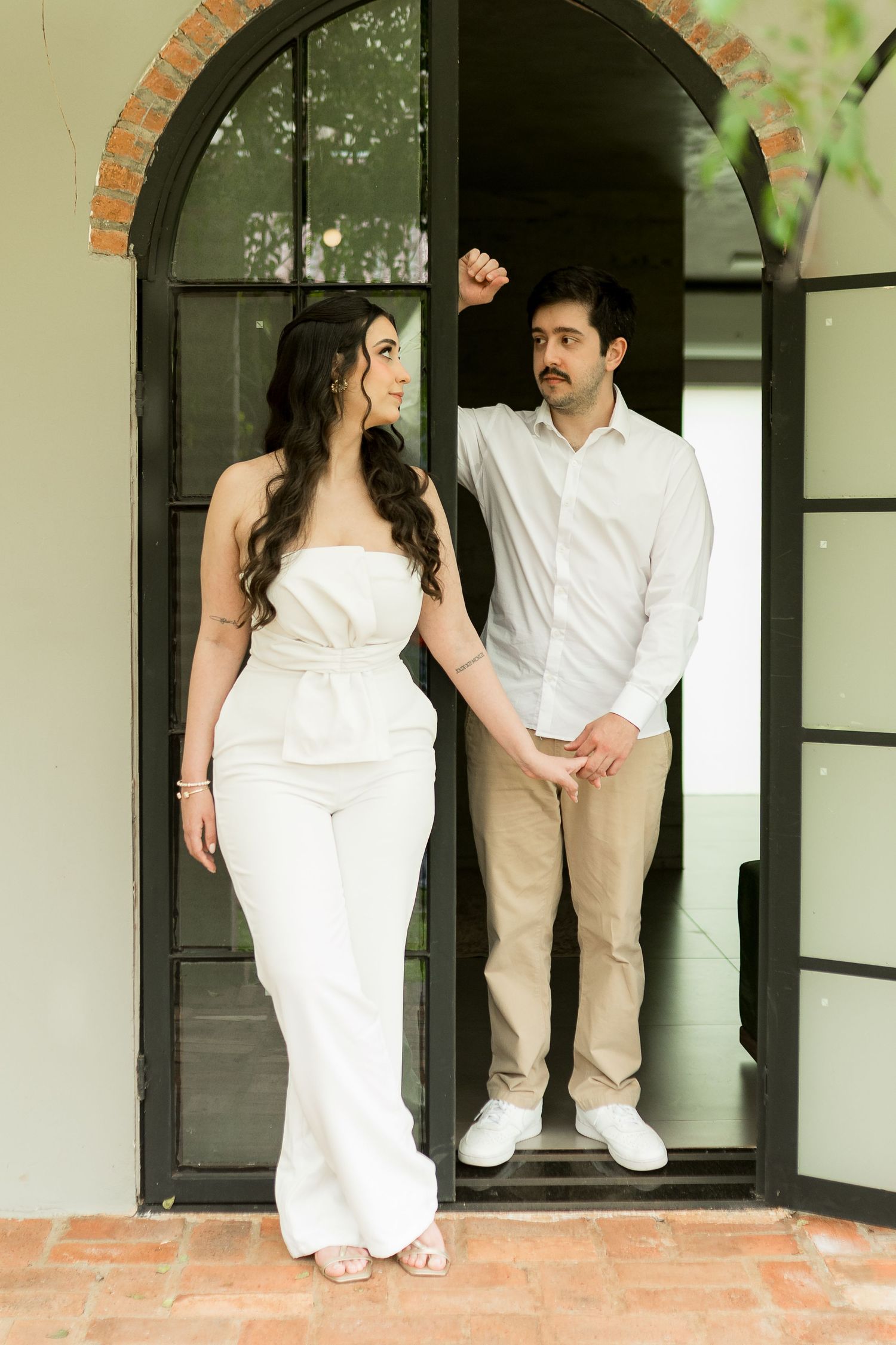 Couple in white outfits holding hands at an arched black door, woman in strapless jumpsuit, man in casual shirt.