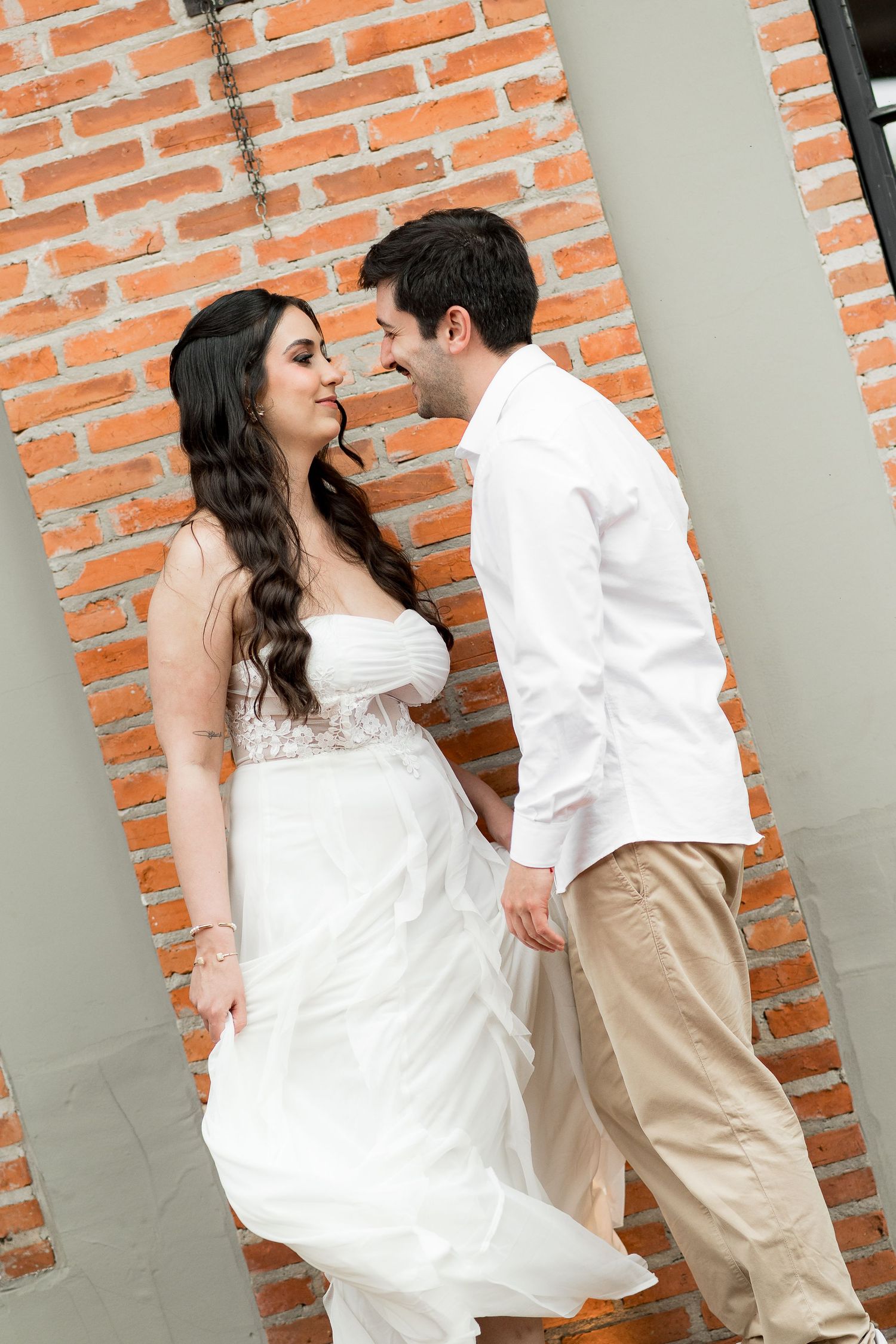 Bride in white gown and groom in white shirt share romantic moment against red brick wall.