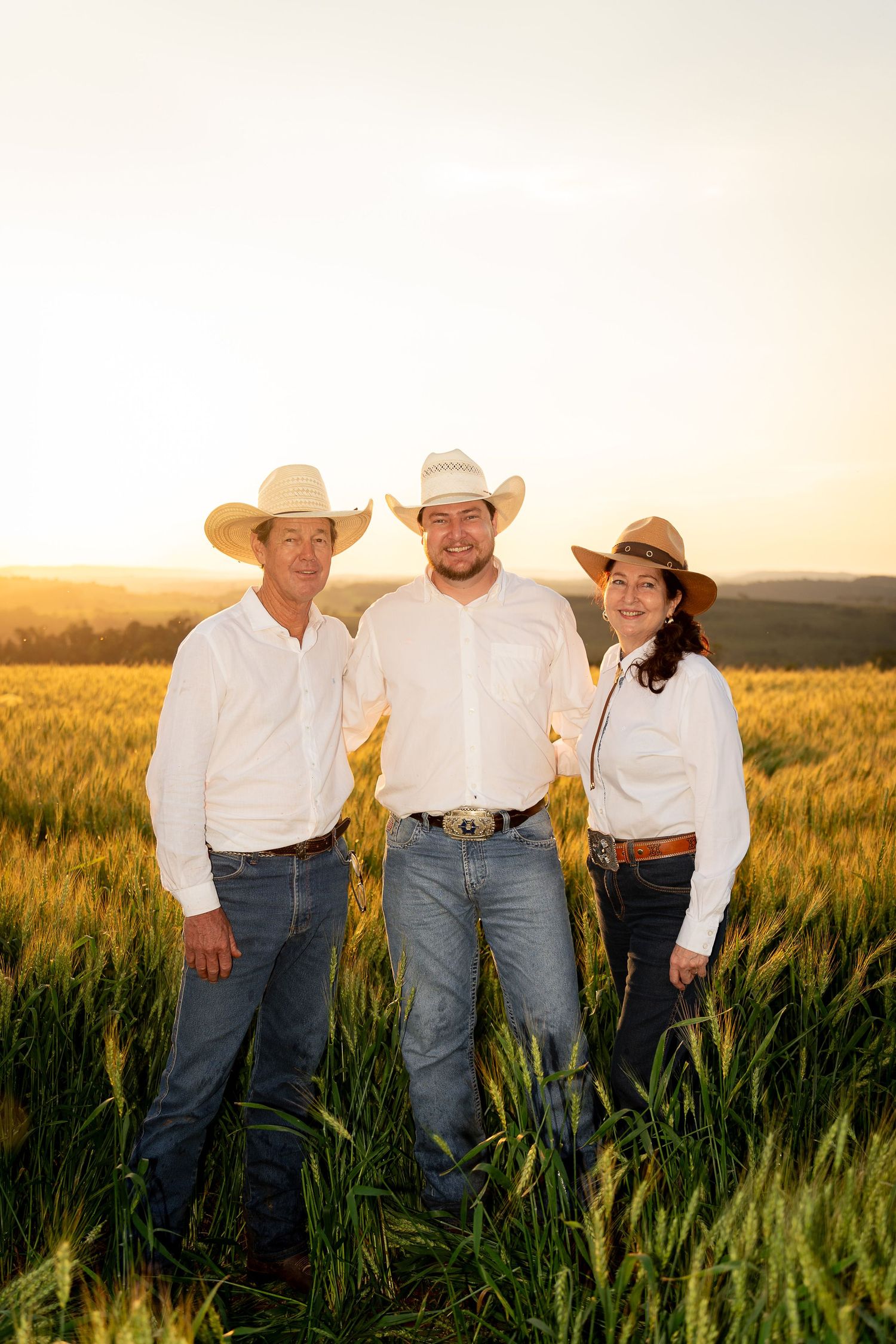 Three people in cowboy hats and white shirts stand smiling in a golden wheat field at sunset.