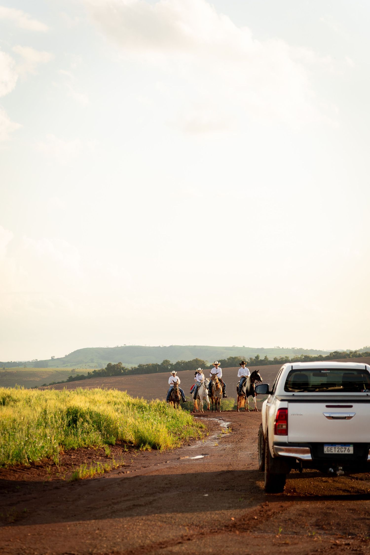 Group of horseback riders on a rural dirt road beside a white pickup truck with vast farmland and hills in background.