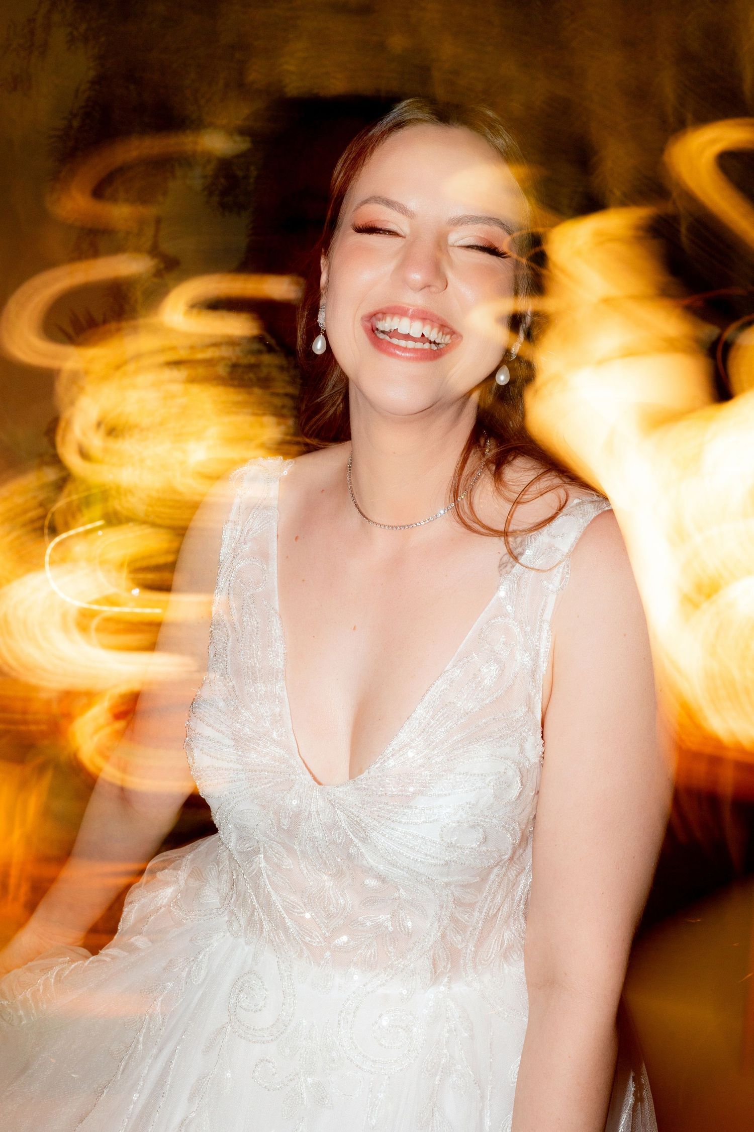 Joyful bride in a white V-neck gown laughs surrounded by swirling golden bokeh light effects.