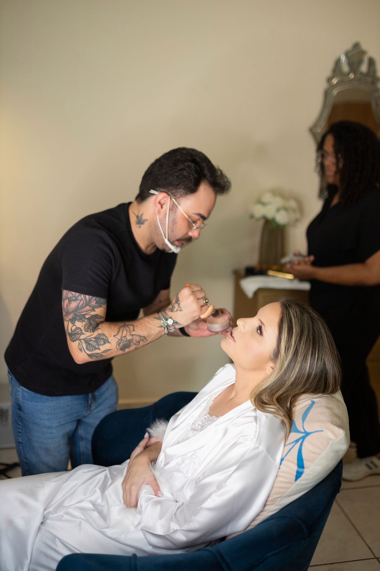 Tattooed makeup artist applying makeup to a woman in a white robe seated in a blue chair at a beauty salon.