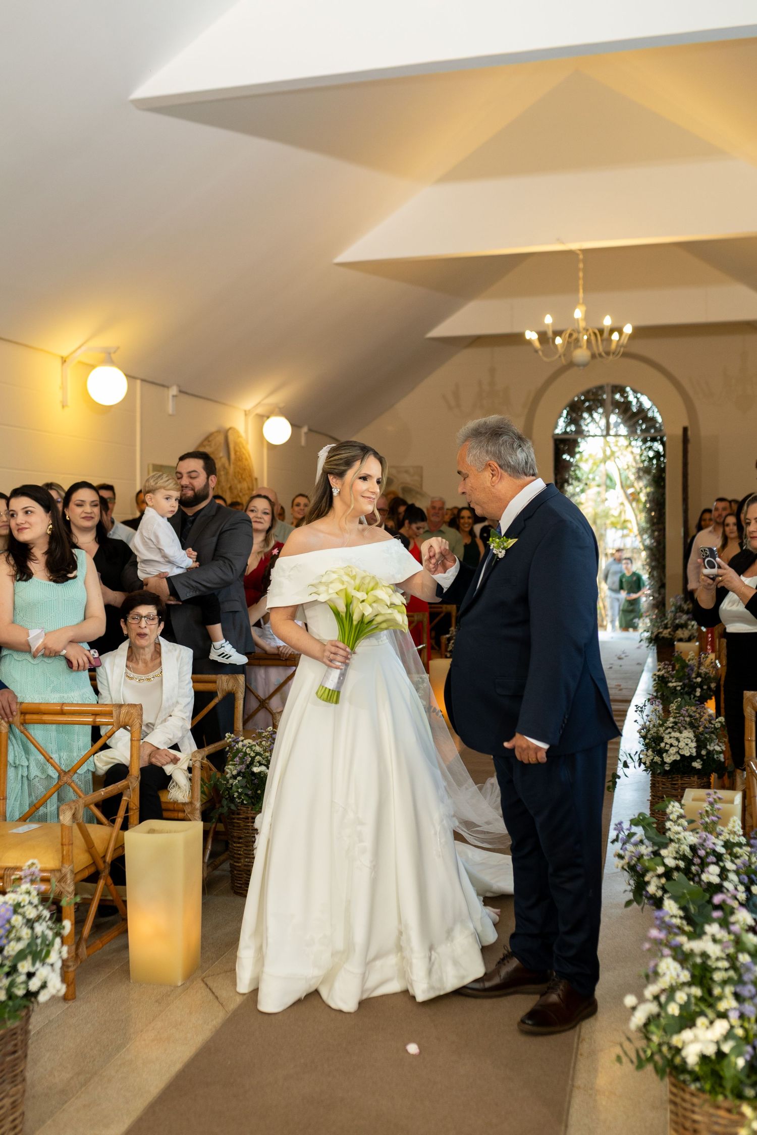 Bride in white off-shoulder gown walks down aisle with escort, holding white bouquet, guests seated in decorated chapel.