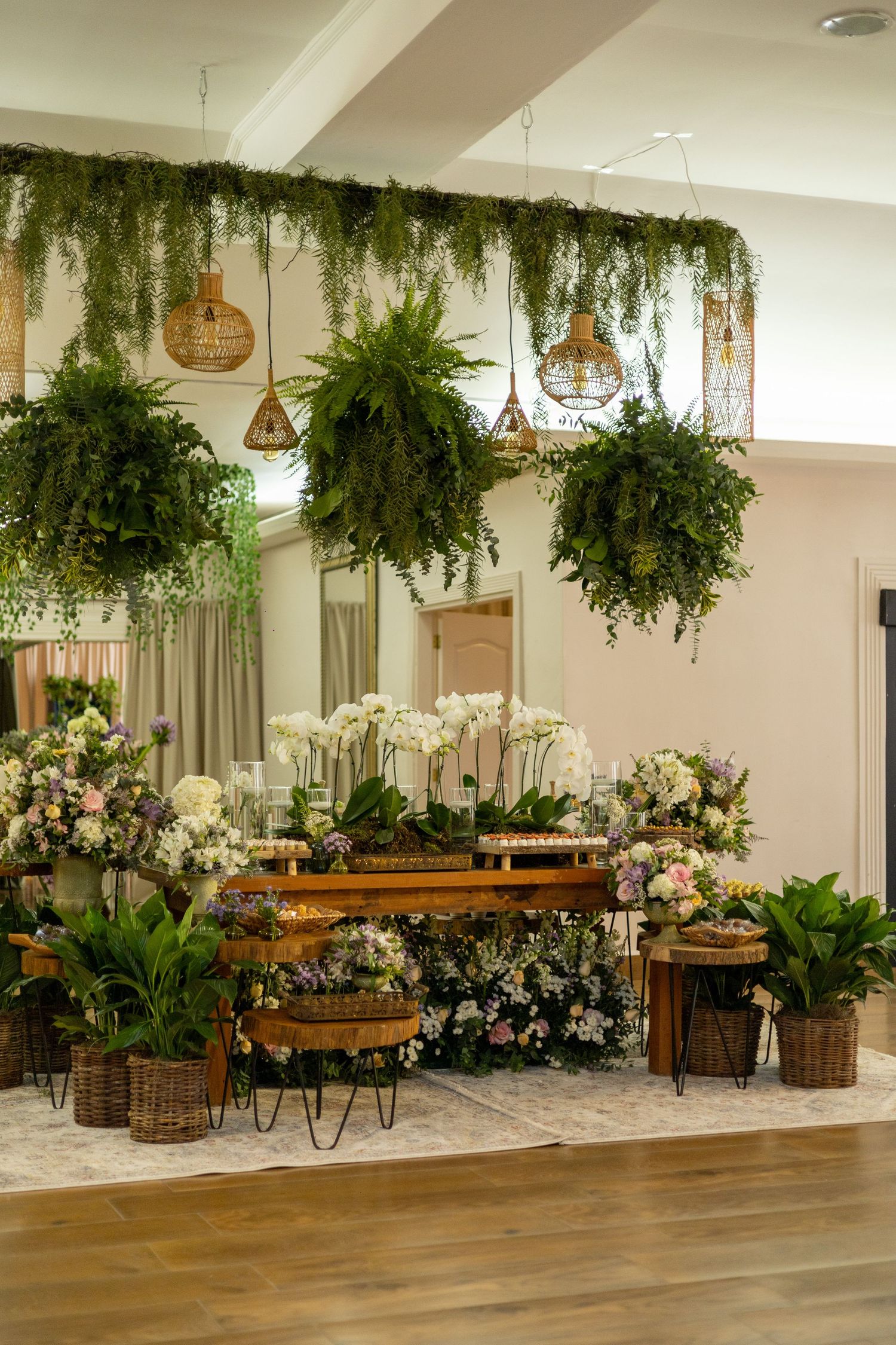 Lush indoor garden display with hanging ferns, orchids, and potted plants arranged on rustic wooden tables.