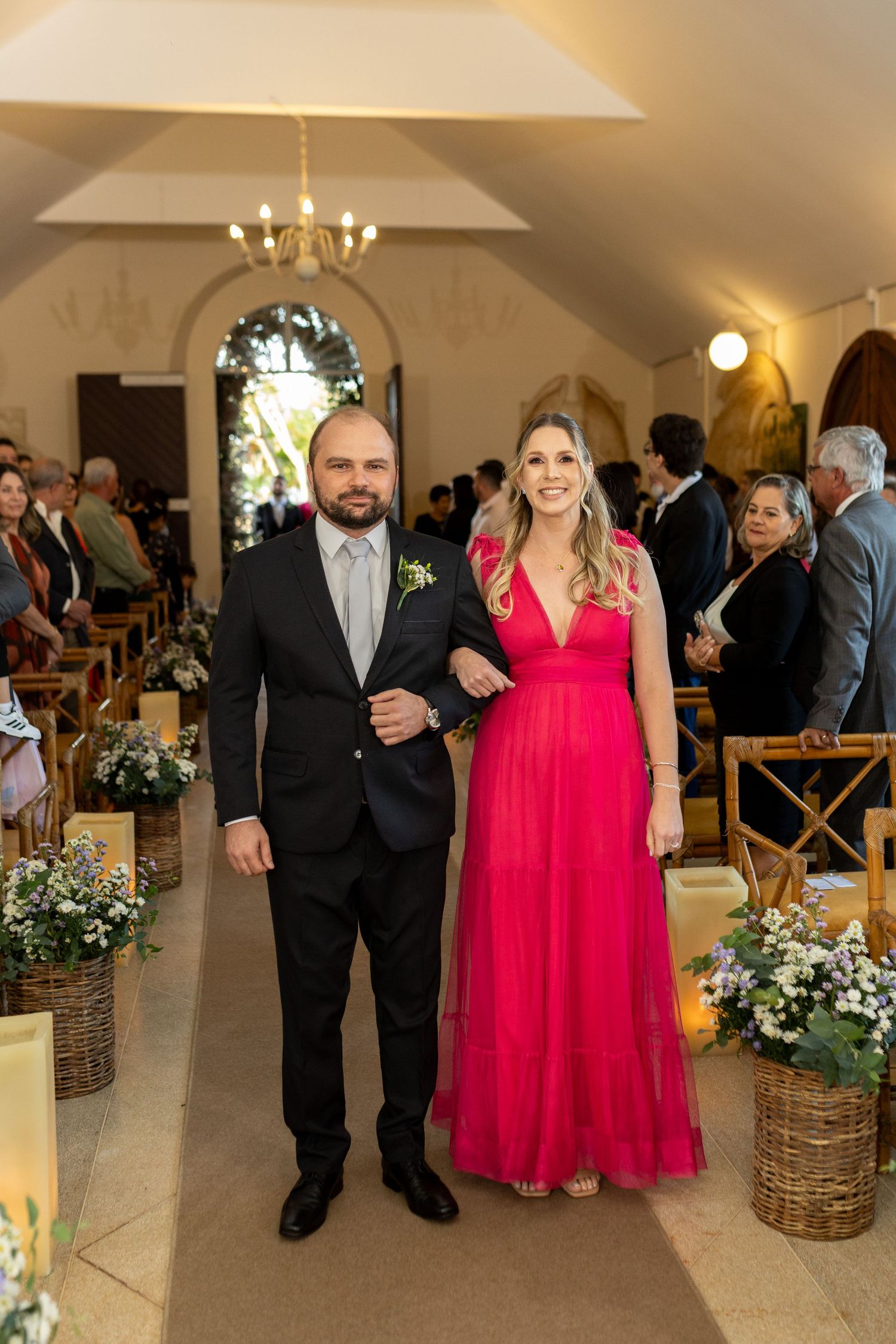 Couple posing in church aisle, man in black suit, woman in red gown, floral decorations, wedding ceremony.