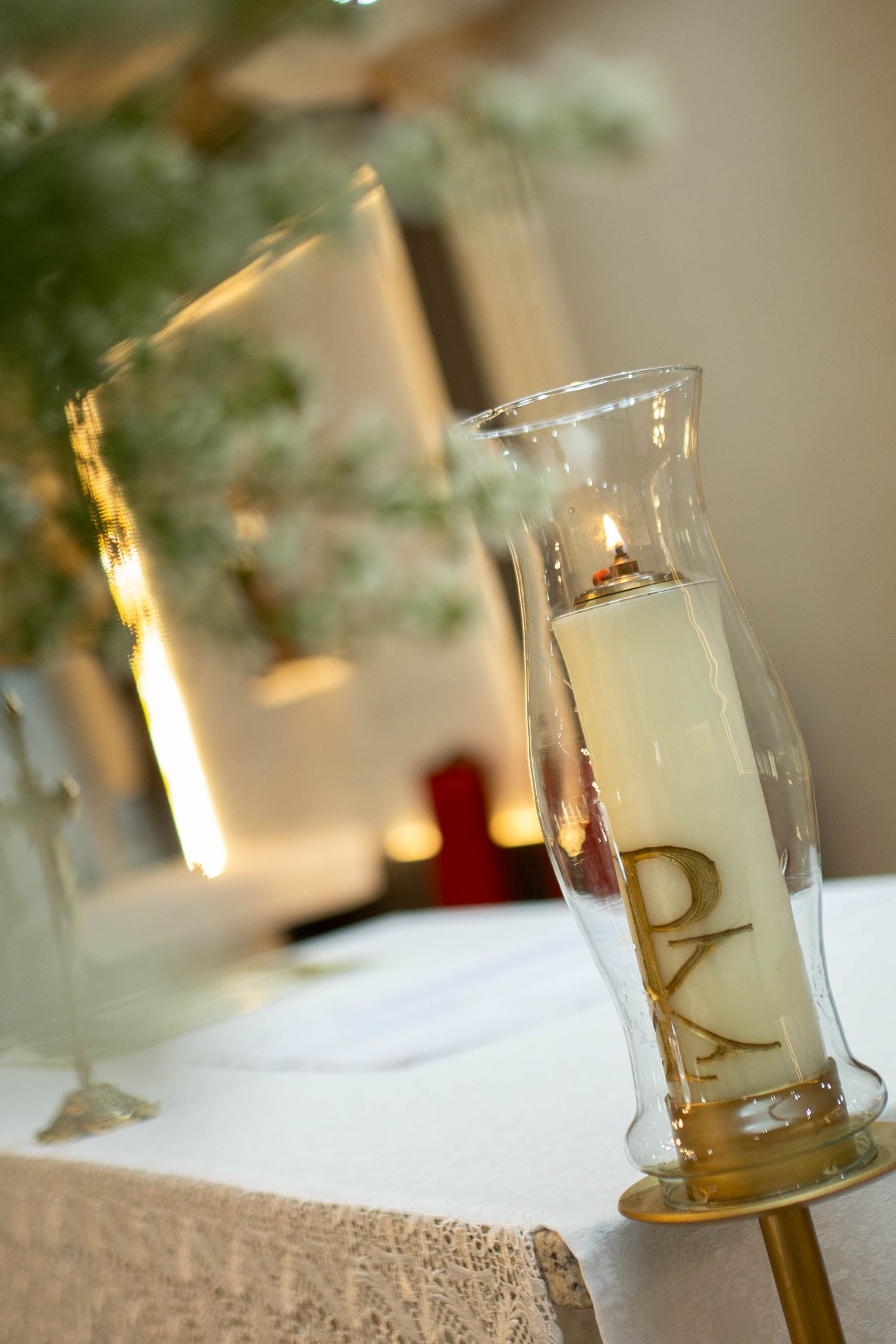Lit Pascal candle with Chi Rho symbol in glass holder on white altar cloth at religious ceremony.