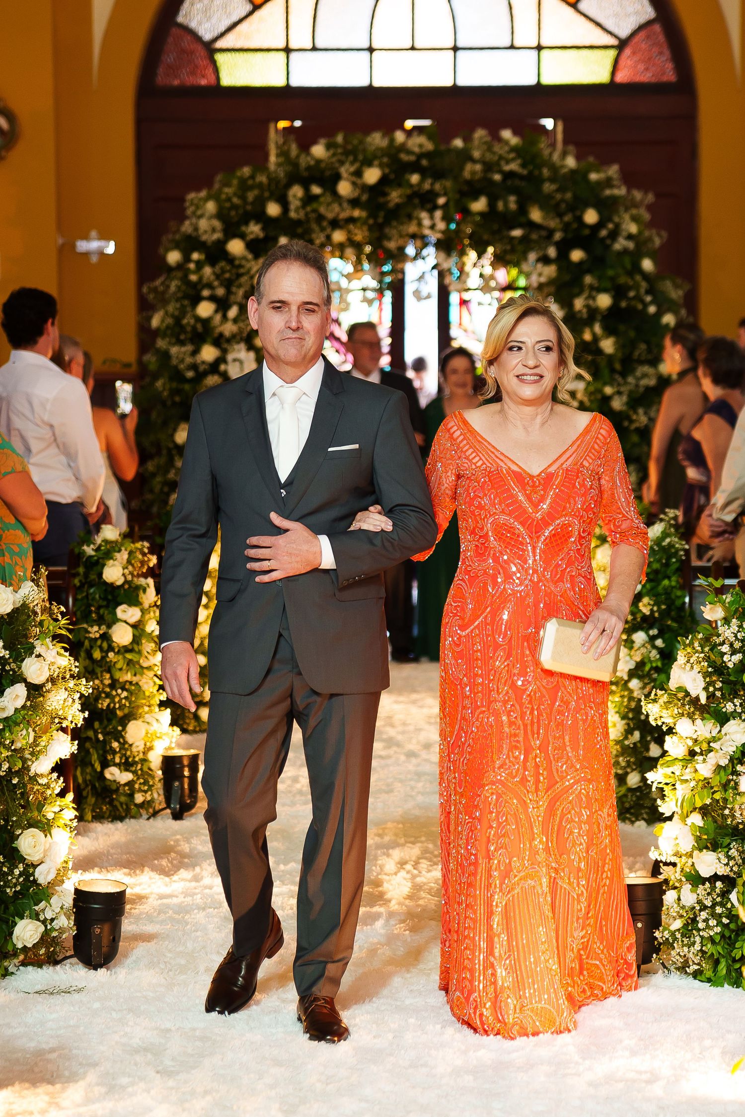Elegant couple walking down floral-decorated wedding aisle, man in dark suit, woman in orange beaded gown.
