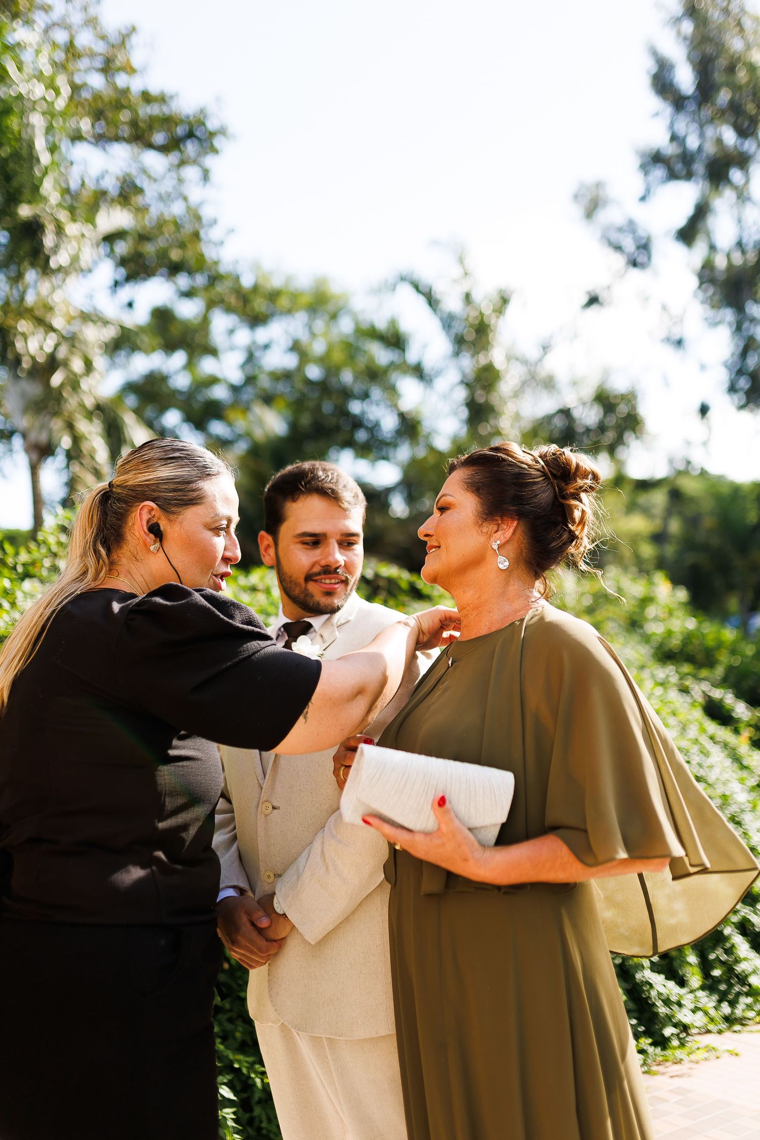 A groom in a white suit stands between two women in black and olive green dresses at an outdoor wedding celebration.