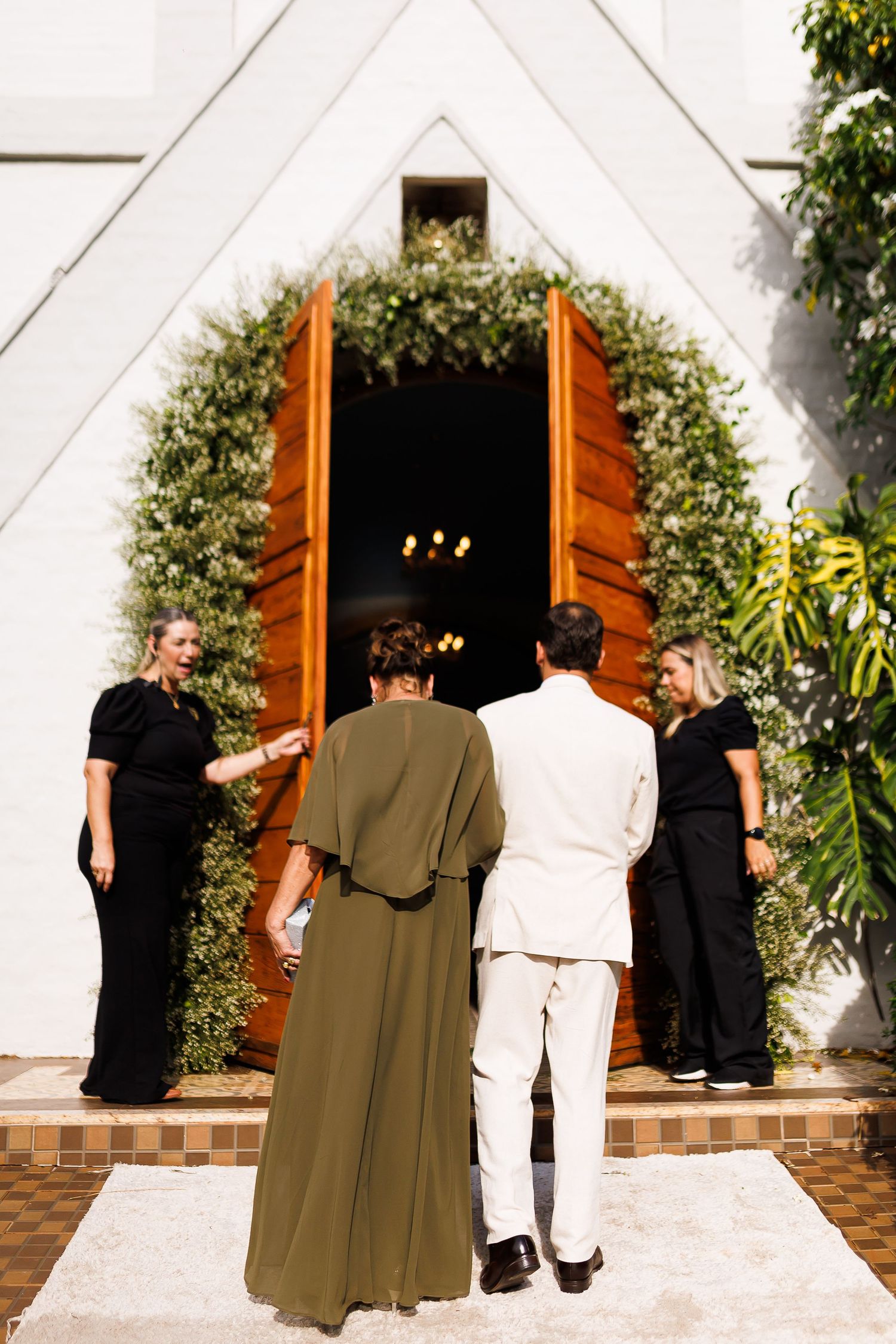 Couple entering a white chapel with wooden doors adorned with greenery, flanked by guests in black attire.