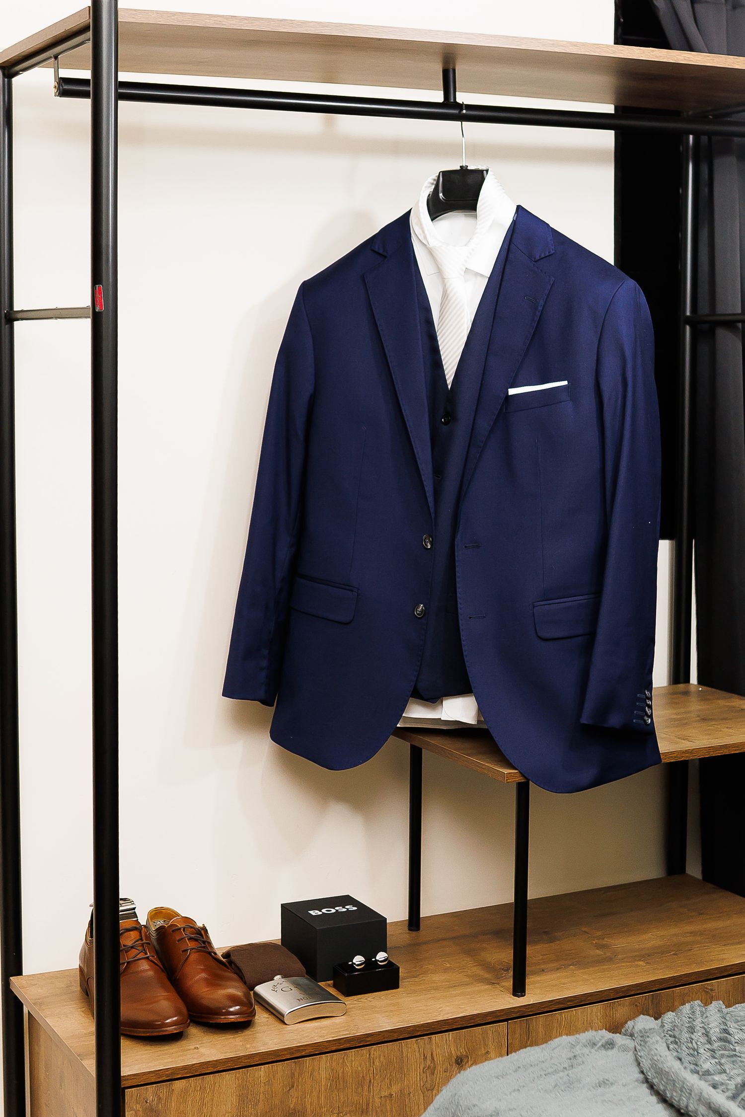 Navy blue three-piece suit displayed on hanger with white dress shirt, brown oxford shoes and accessories on wooden shelf below.