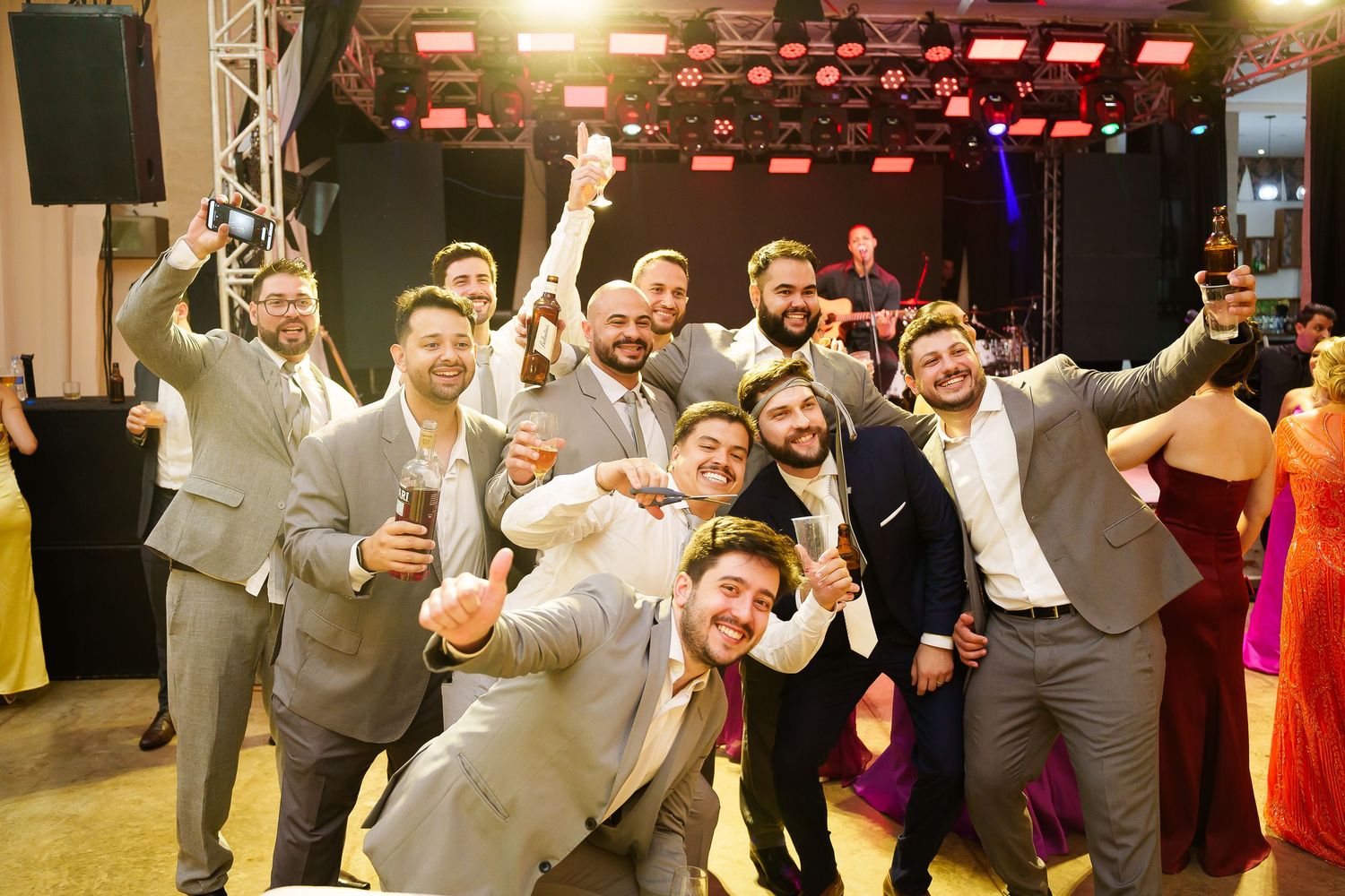 Group of groomsmen in gray suits celebrating at a wedding reception, raising glasses and posing for a fun photo.