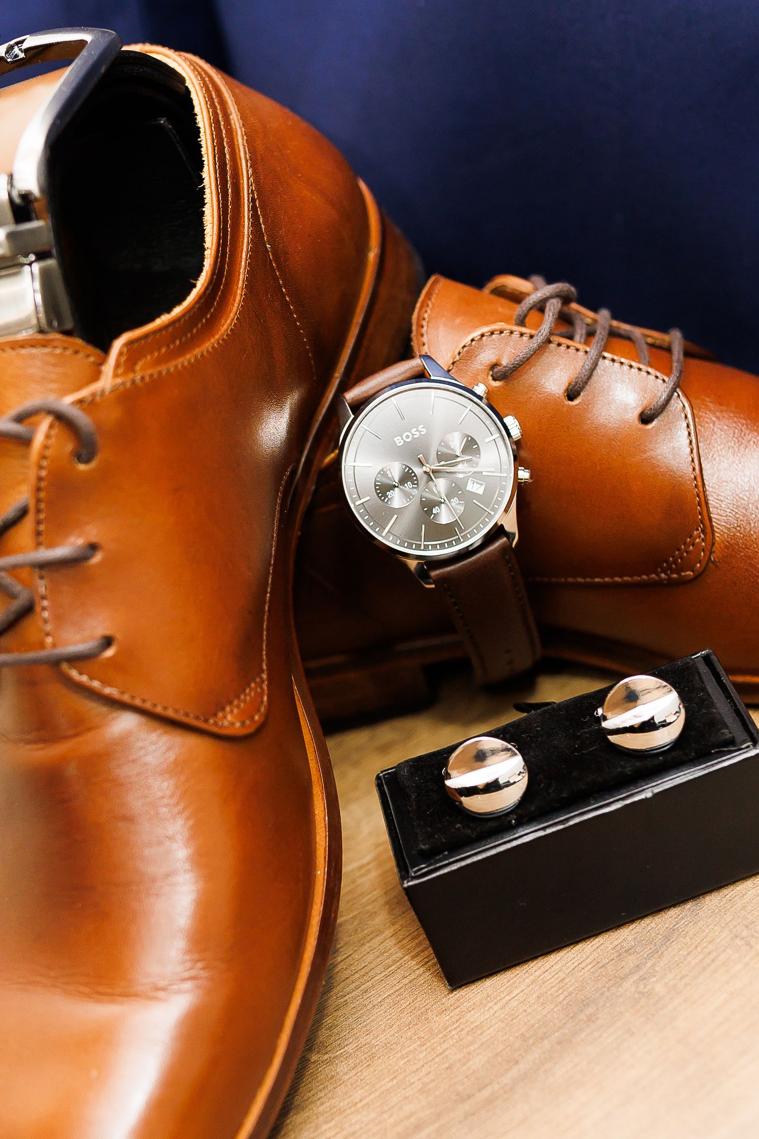 Tan leather Oxford dress shoes displayed with a silver chronograph watch and cufflinks on a wooden surface.