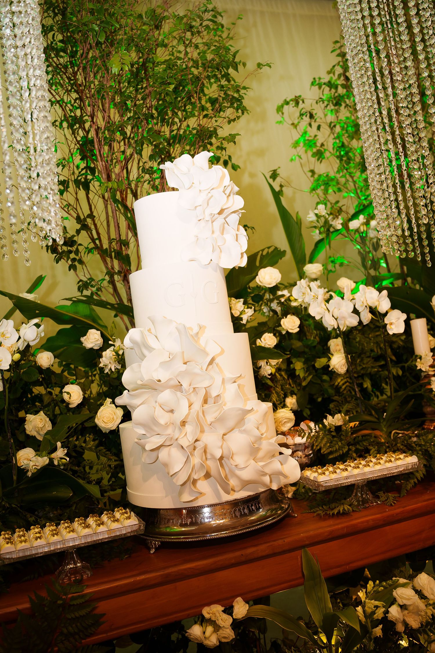 Elegant three-tier white wedding cake adorned with large fondant flowers, displayed among lush greenery and white roses.