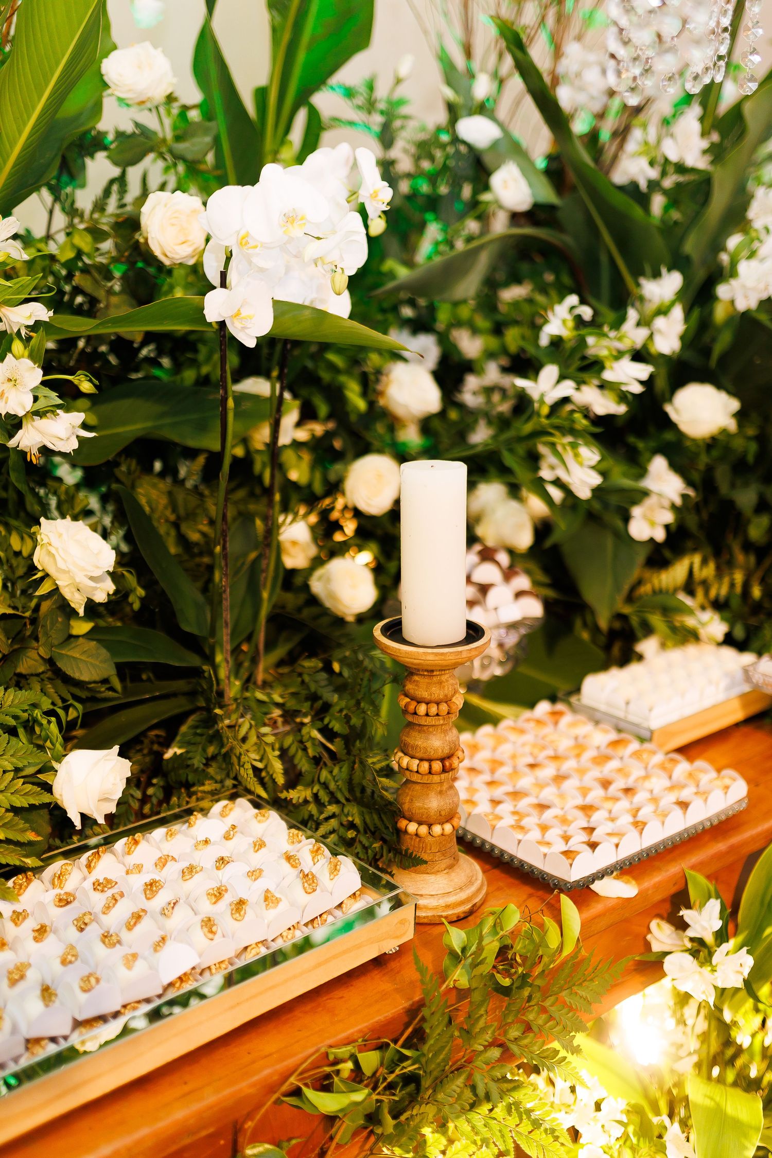 Elegant wedding dessert table with white candle, orchids, roses, and assorted sweets amid lush green tropical foliage.