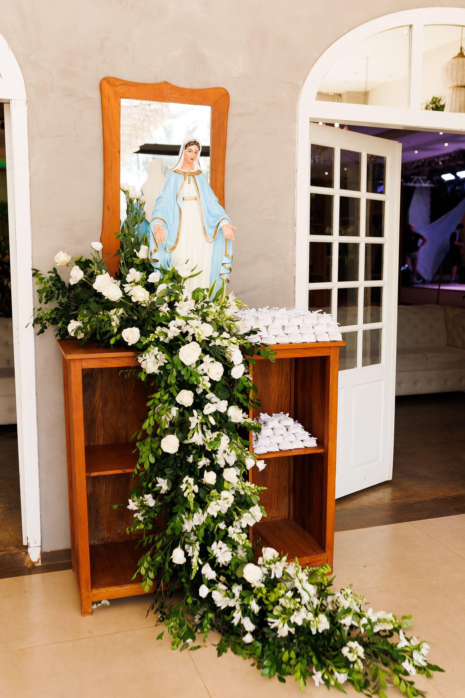 Wooden shelf decorated with white flowers and greenery garland beside a Virgin Mary figurine for a religious celebration.