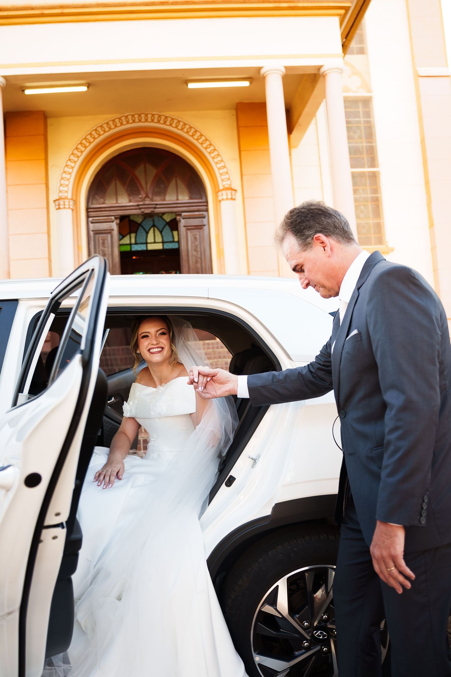 A smiling bride in a white gown exits a white luxury car assisted by a man in a suit outside a church.