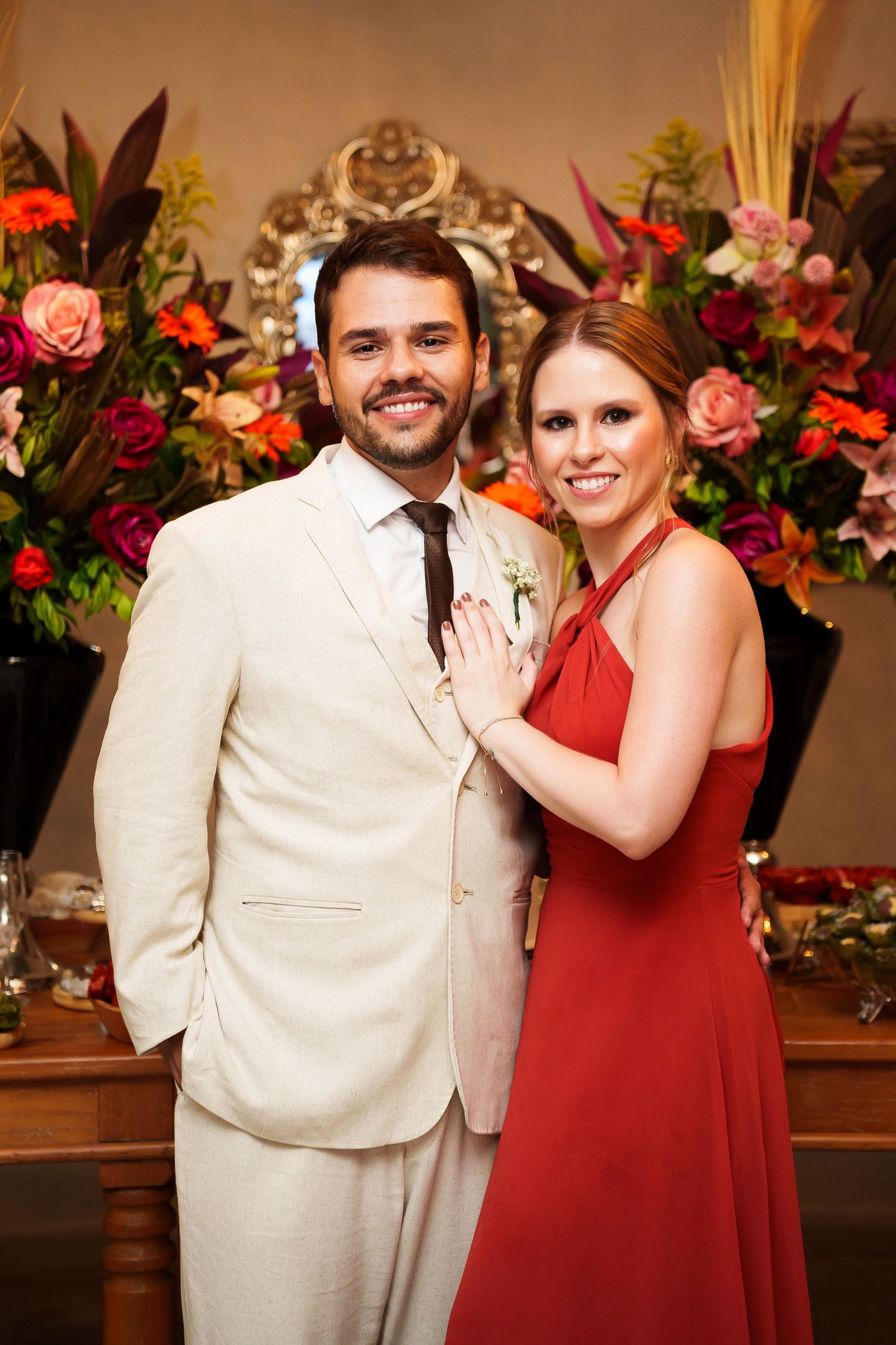 Smiling couple posing at a wedding, man in cream suit, woman in red dress, colorful floral arrangement in background.