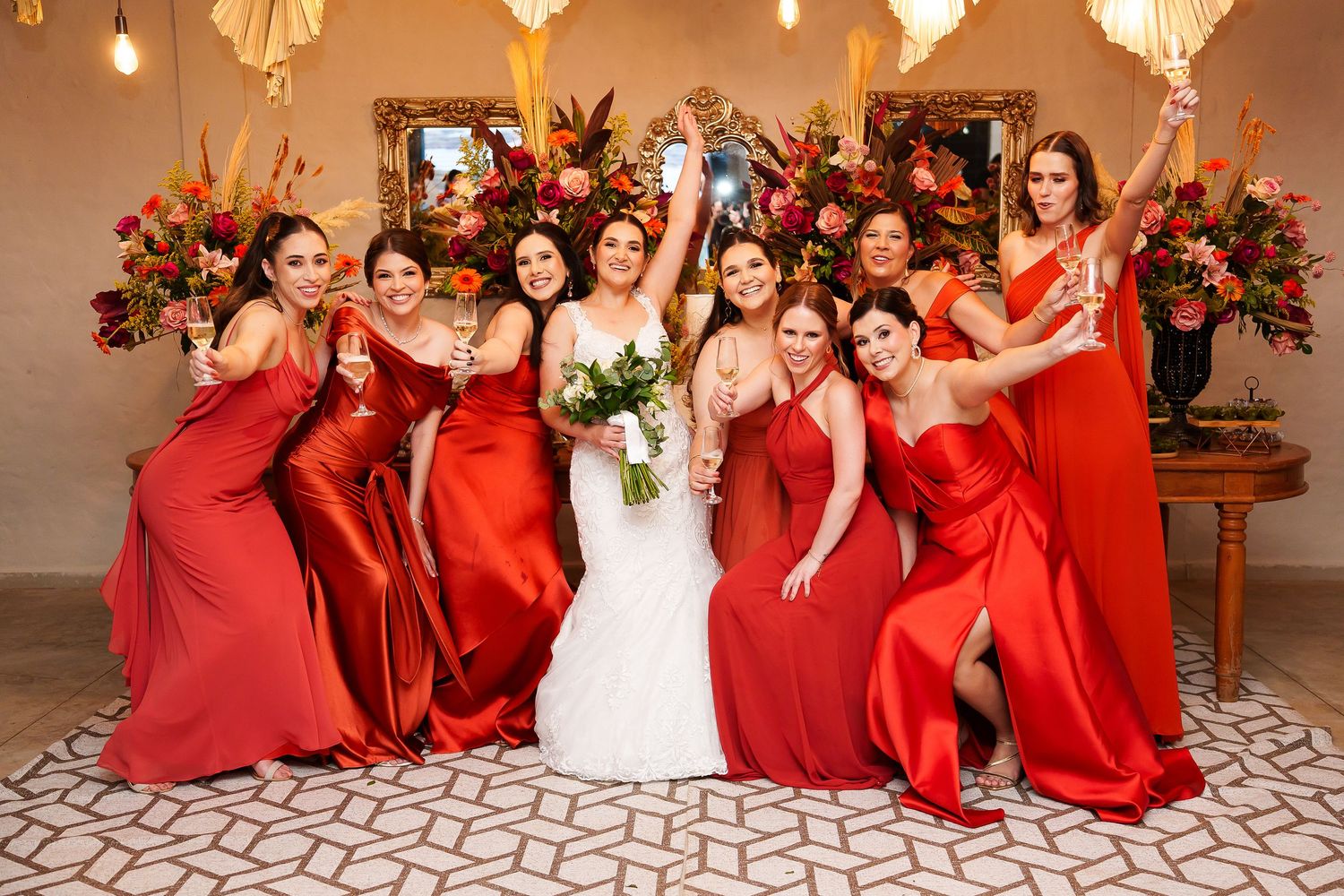 Bride in white gown poses with eight bridesmaids in red satin dresses, holding bouquets and champagne at a wedding reception.