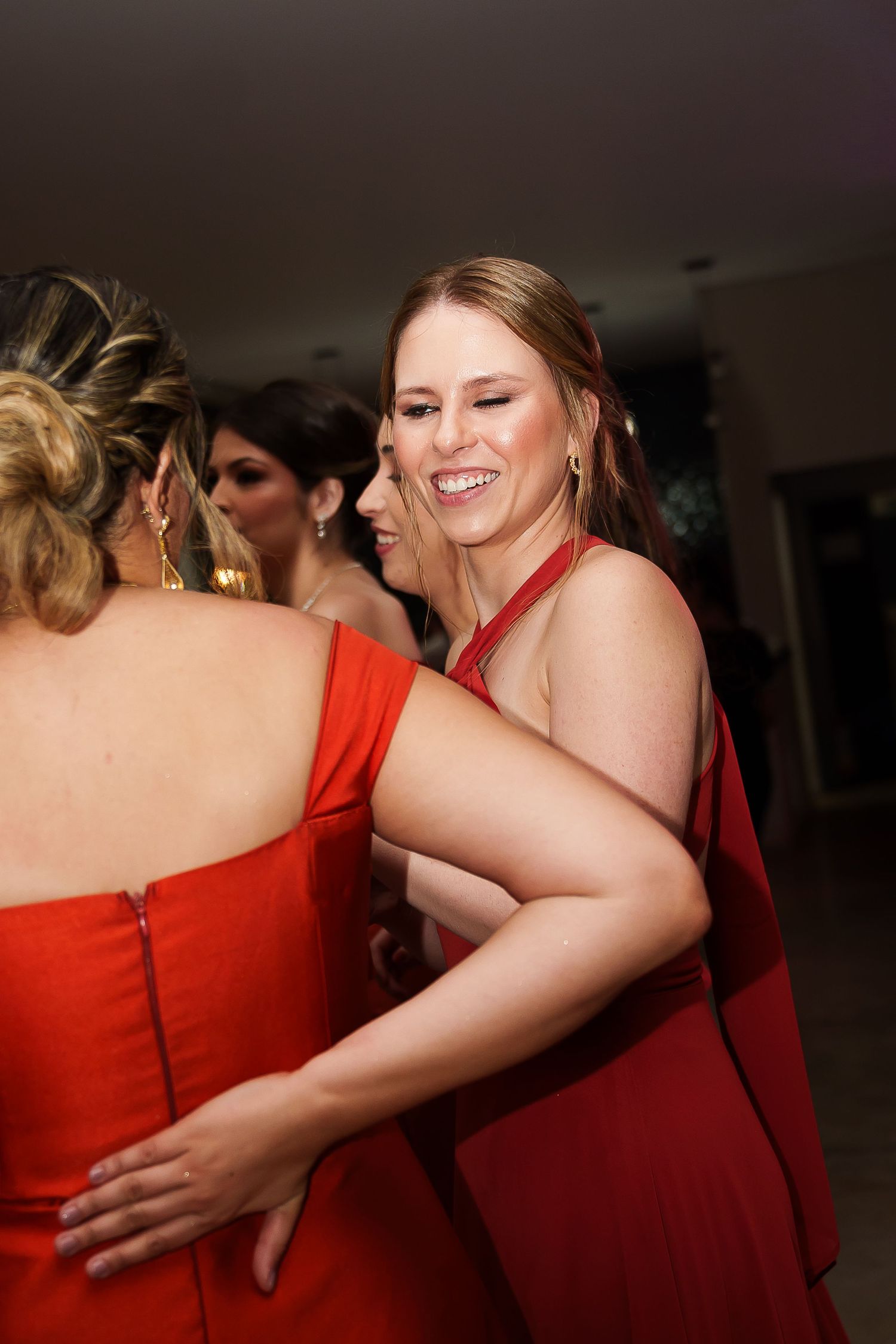 Two women in red dresses share a warm hug and smile at a formal event or party.