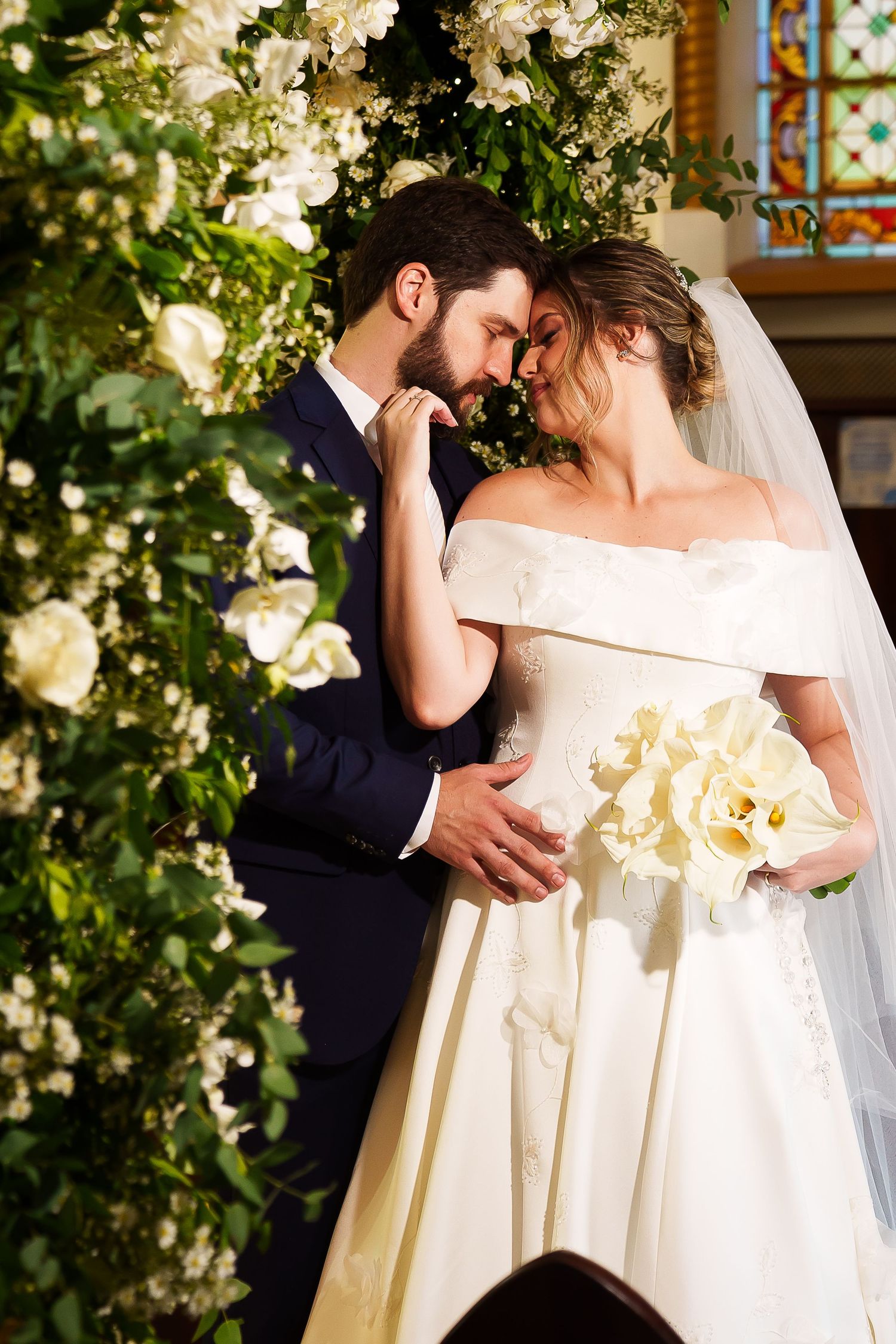 Bride and groom share romantic moment at church altar surrounded by white floral arrangements, bride holding calla lily bouquet.