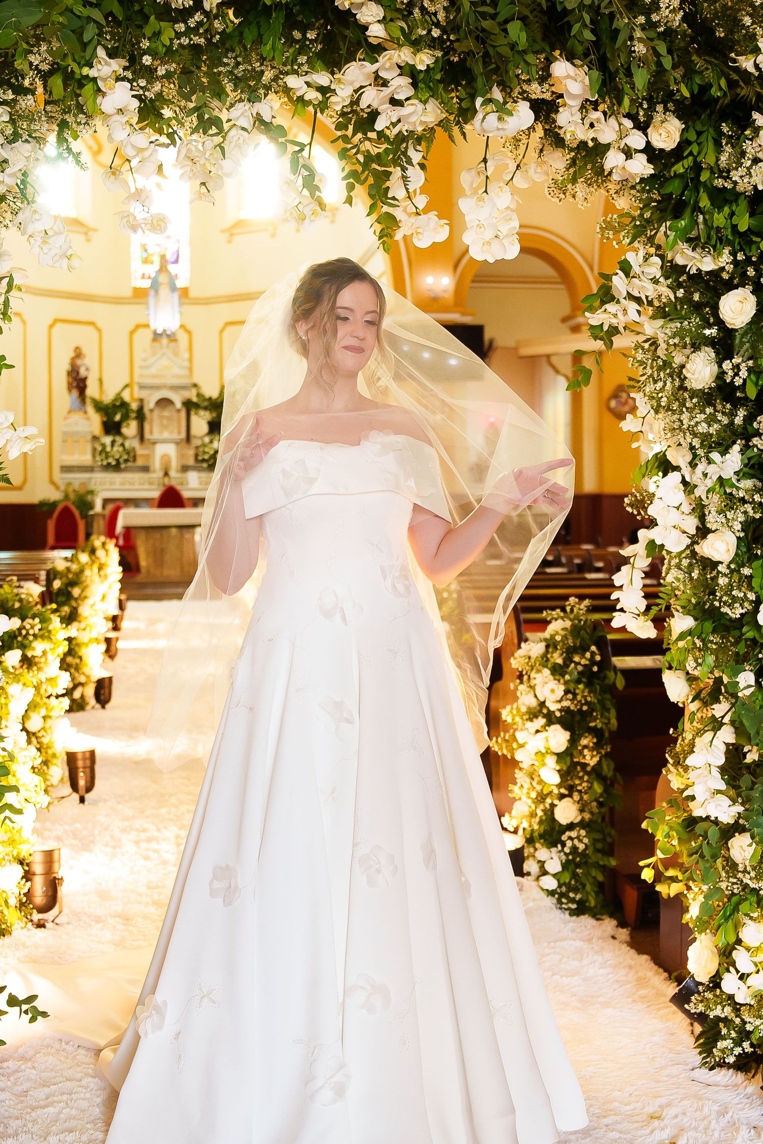 Bride in off-shoulder white ball gown with veil stands under floral arch in elegantly decorated church.