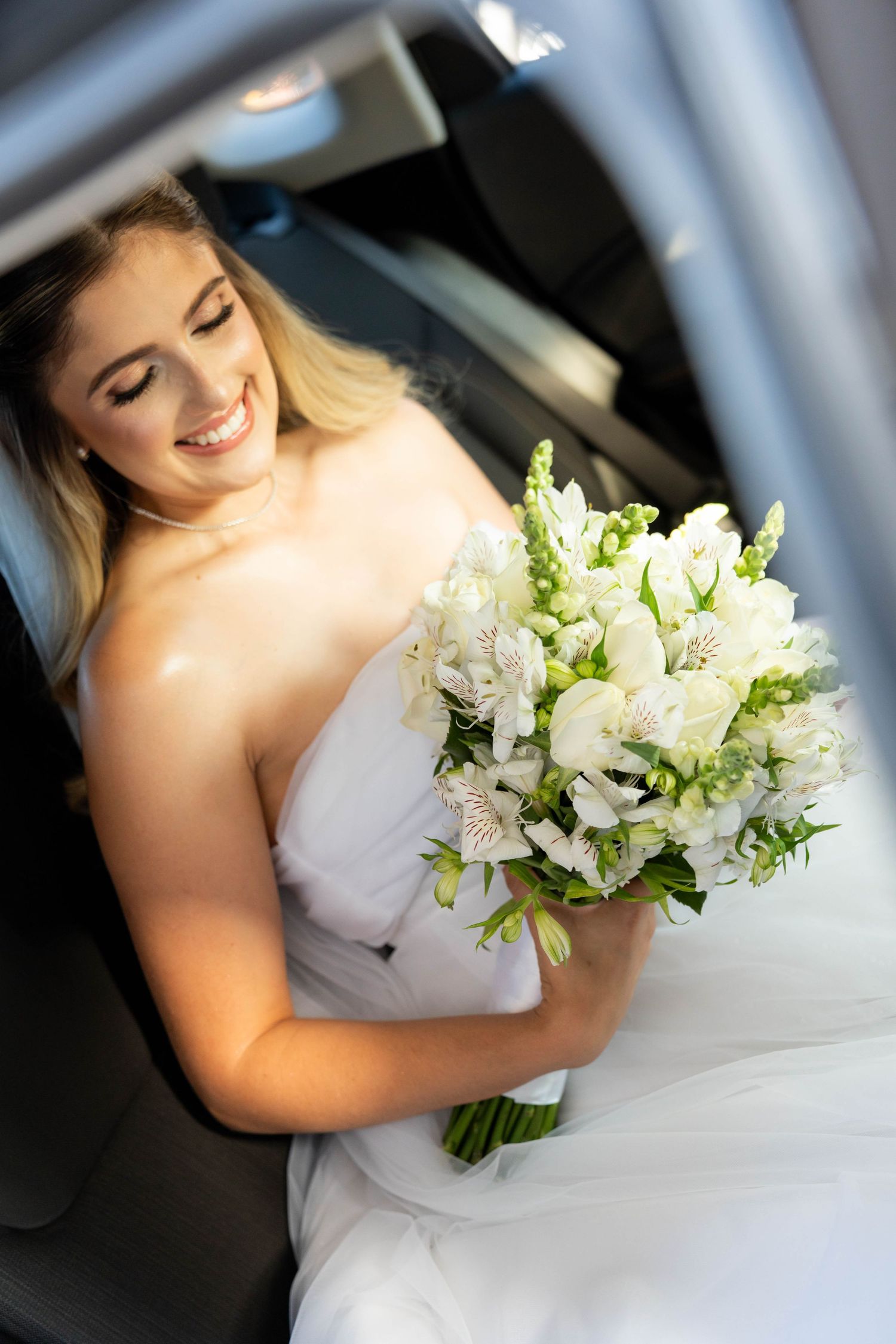 Smiling bride in white strapless gown holding elegant white floral bouquet while sitting in a car on her wedding day.