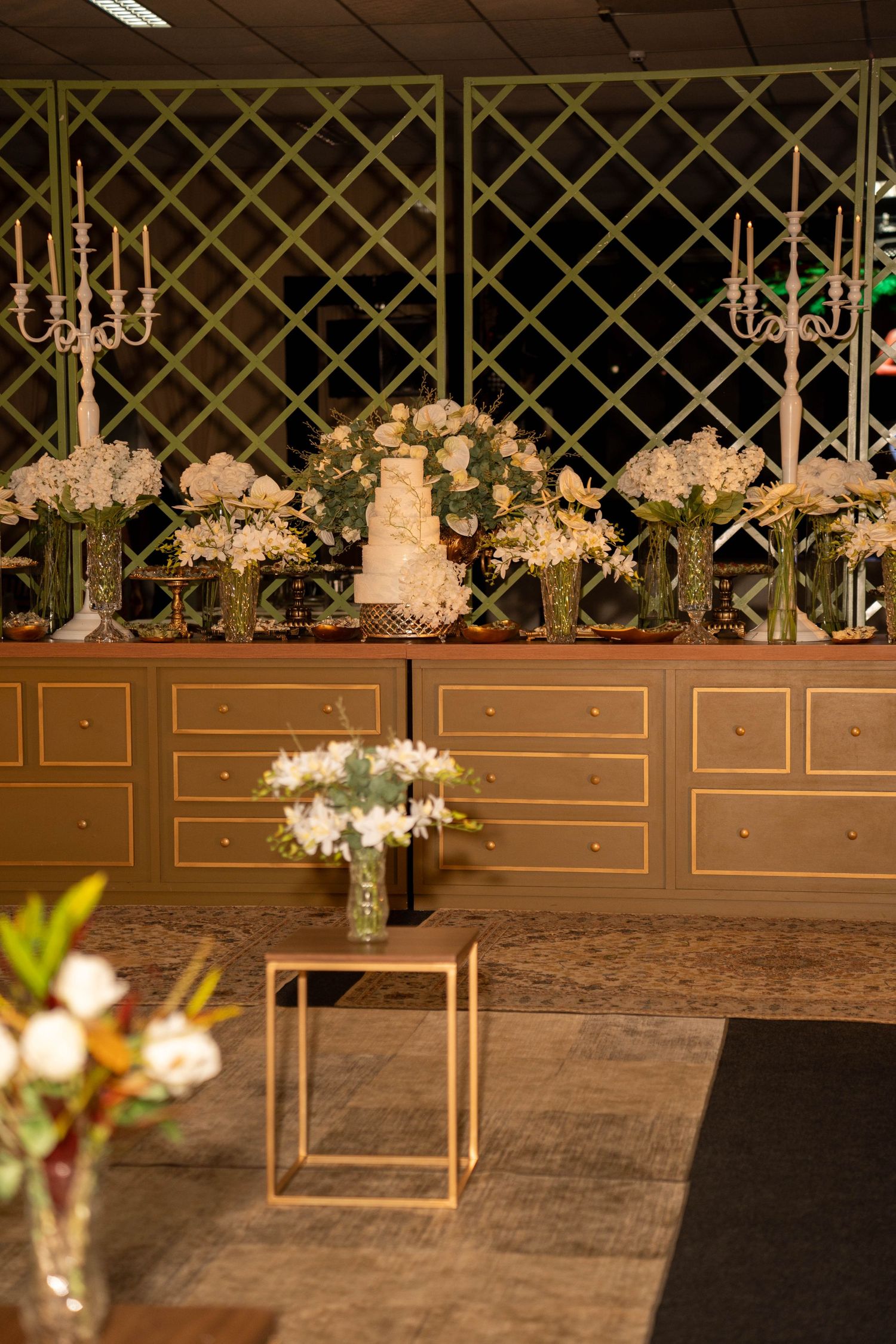 Elegant wedding cake display on gold dresser with white floral arrangements and candelabras against lattice backdrop.