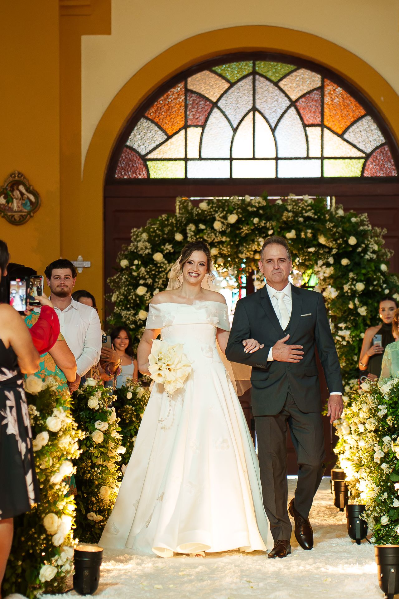Bride in white gown walks down church aisle with escort, framed by floral arch and stained glass window.