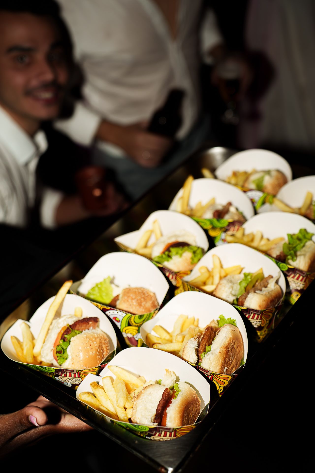 Waiter serving a tray of mini burgers and french fries in individual portions at a catered event.