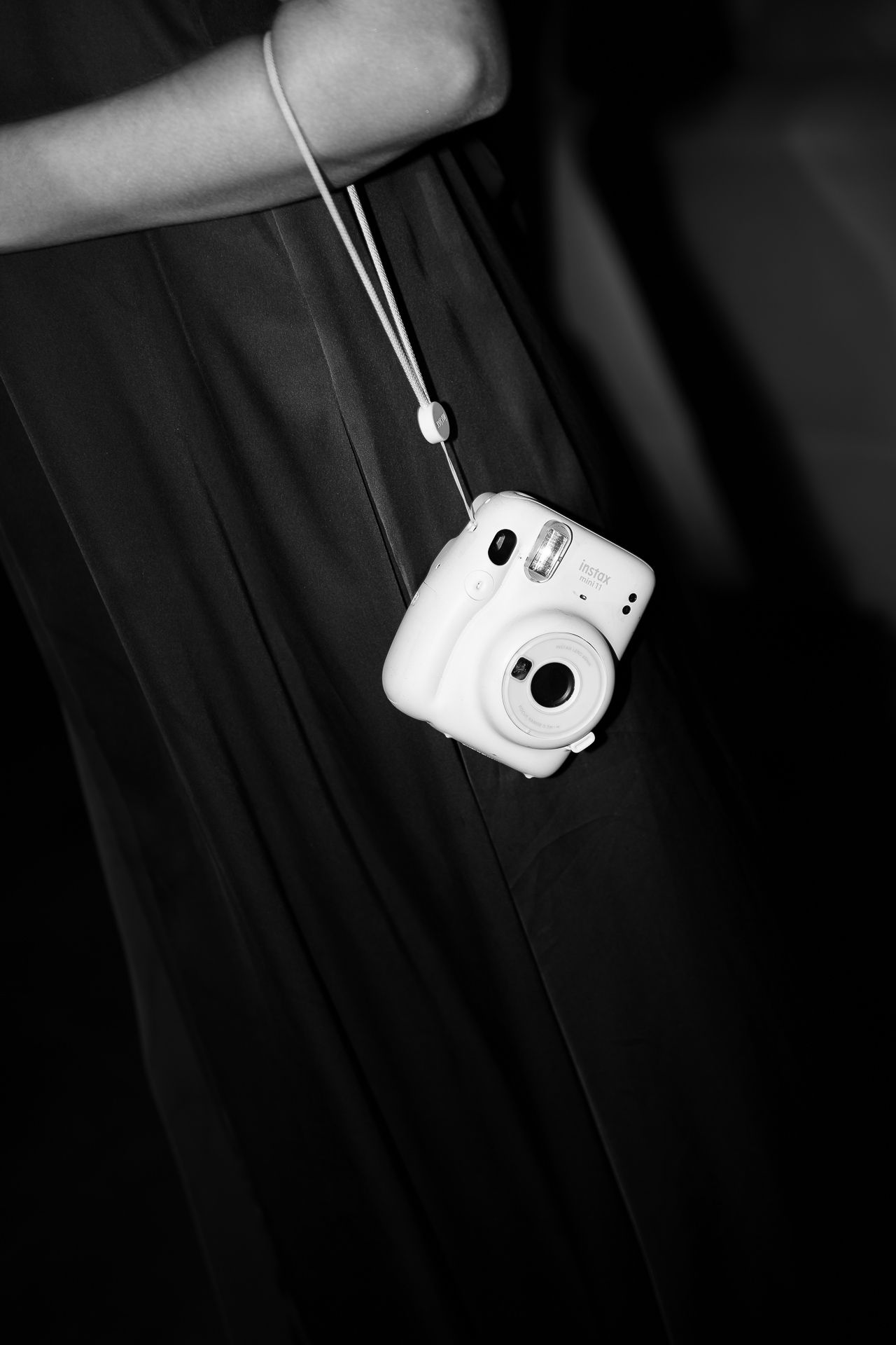 Black and white photo of a person holding a white Fujifilm Instax Mini camera on a strap against a dark skirt.
