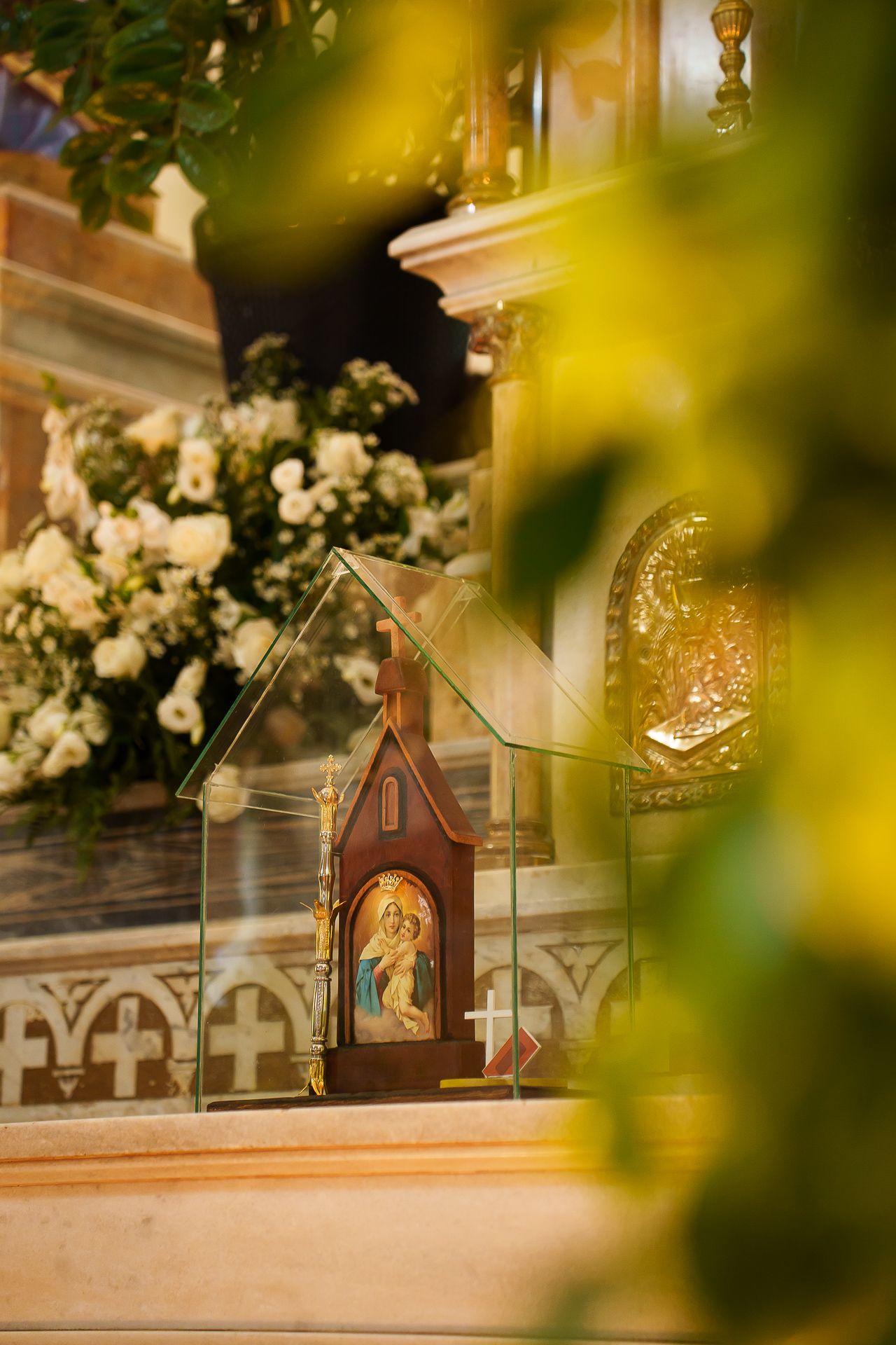 Ornate religious icon in wooden shrine displayed on church altar surrounded by white floral arrangements.