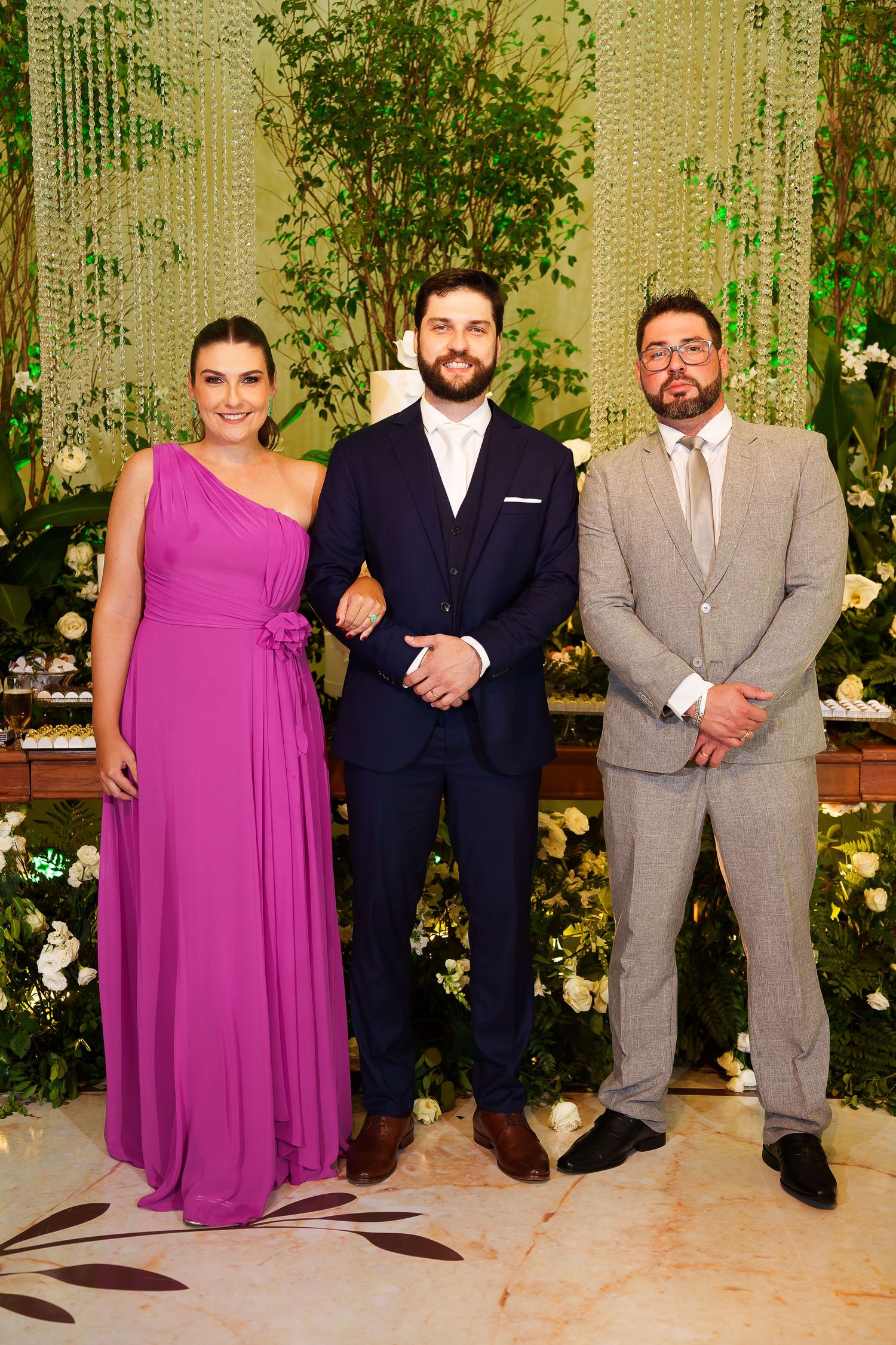 Three elegantly dressed guests pose together at a formal wedding reception with floral decorations.