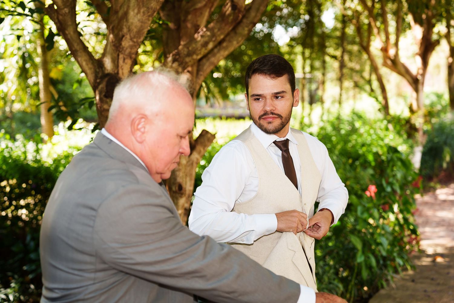 Groom in white shirt and tan vest stands outdoors near trees at wedding ceremony with older man beside him.