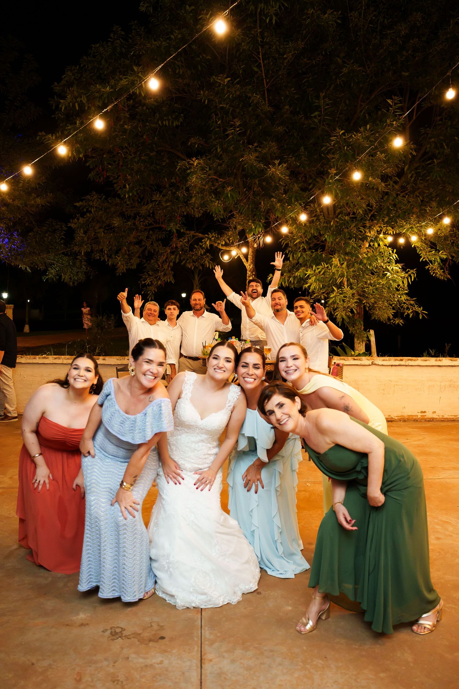 Bride poses with bridesmaids in colorful gowns outdoors at night wedding reception with string lights and cheering guests.