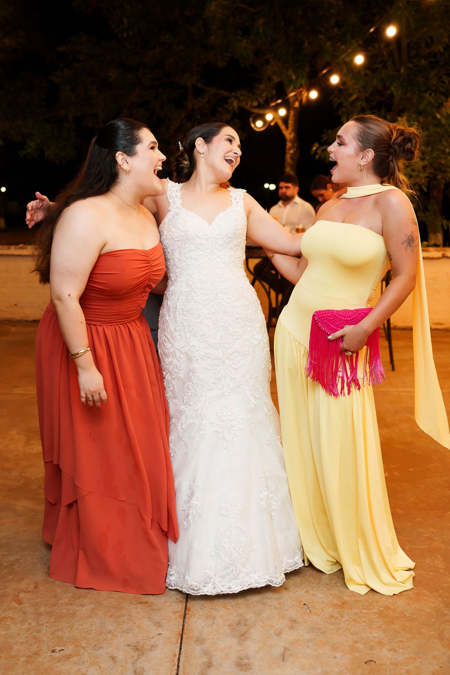 Bride in white lace gown laughing with two bridesmaids in orange and yellow dresses at outdoor wedding reception.