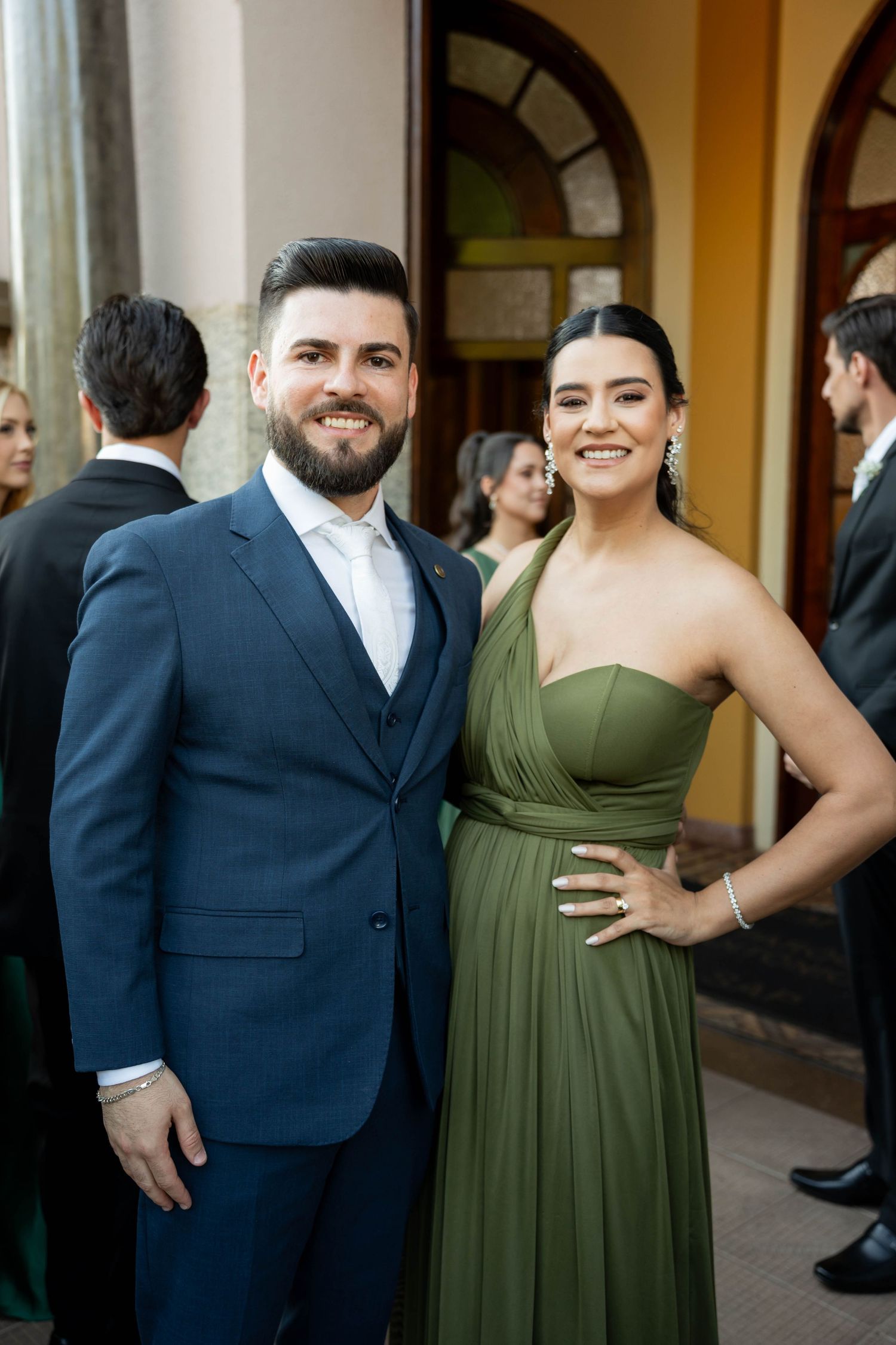 A smiling couple poses outdoors, man in navy suit and woman in elegant olive green one-shoulder gown at a formal event.