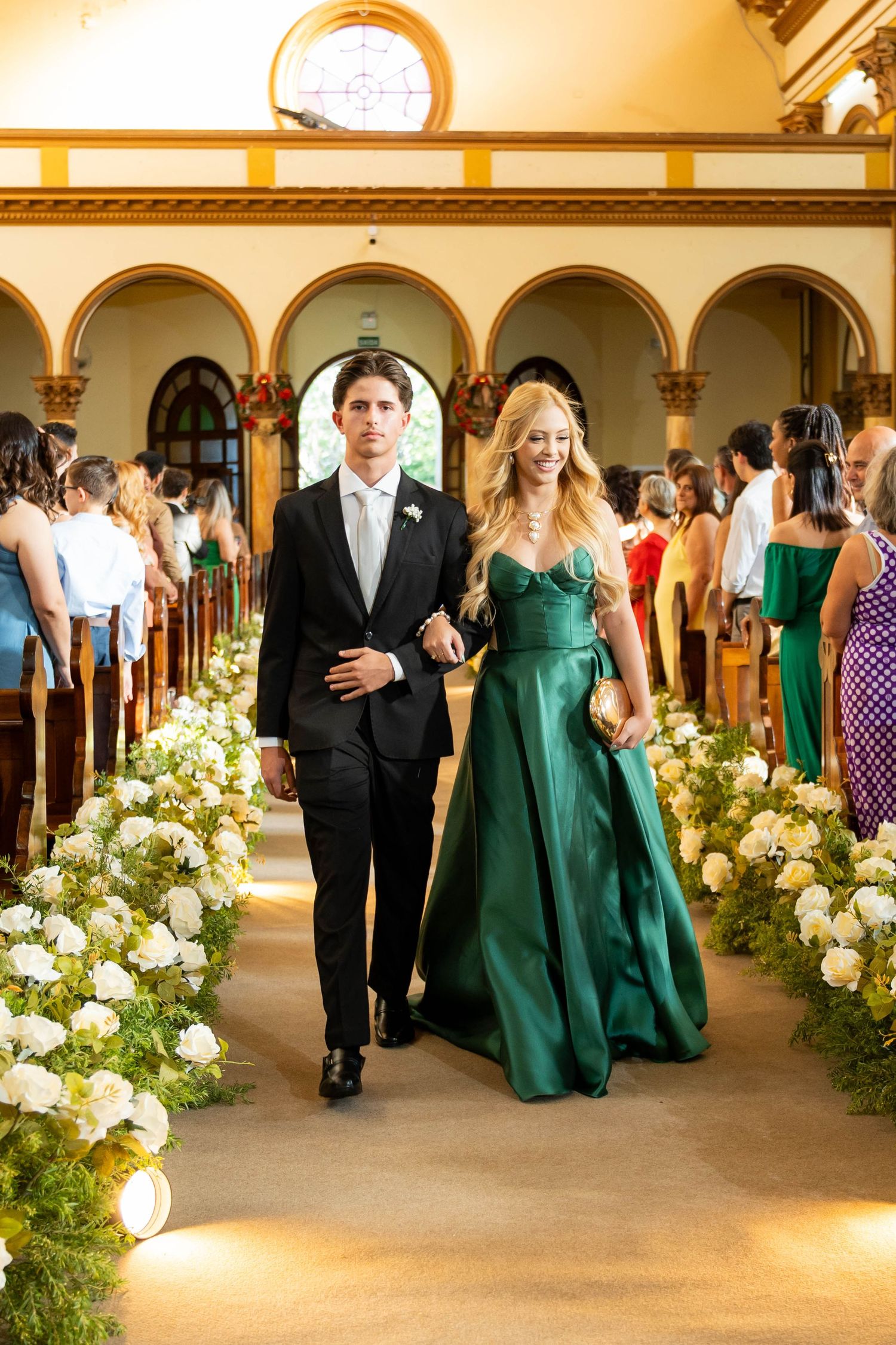 A man in a black tuxedo and woman in an emerald green gown walk down a flower-lined church aisle at a wedding.