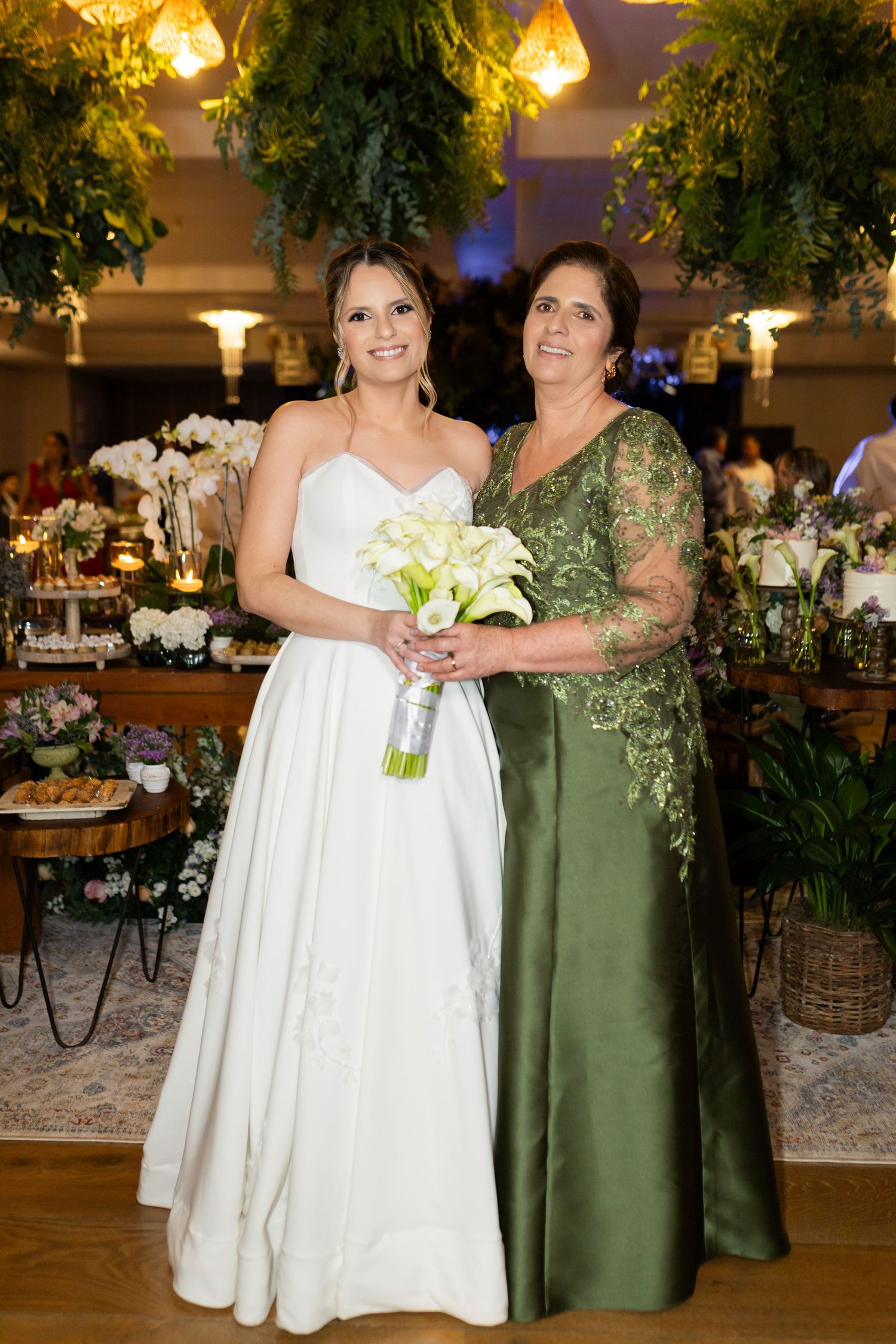 Bride in white gown holds calla lily bouquet beside woman in green lace dress at elegant floral wedding reception.