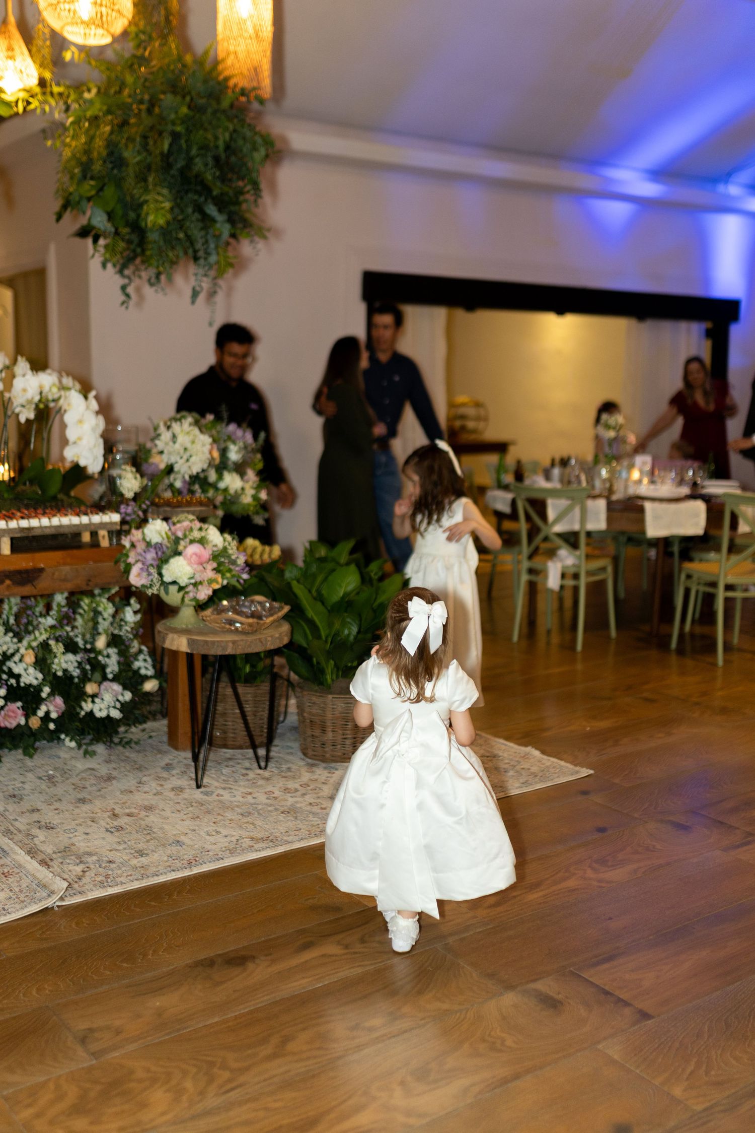 Young flower girl in white dress with bow walks across wooden dance floor at elegant wedding reception.