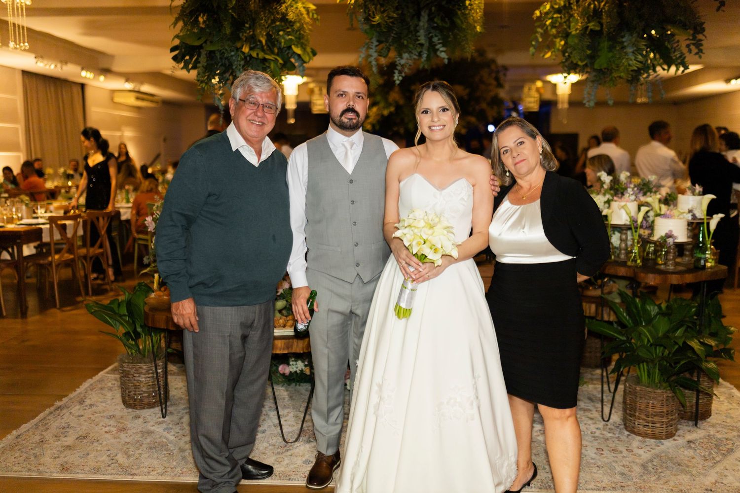 Bride and groom pose with two family members at an elegant indoor wedding reception with lush greenery decor.