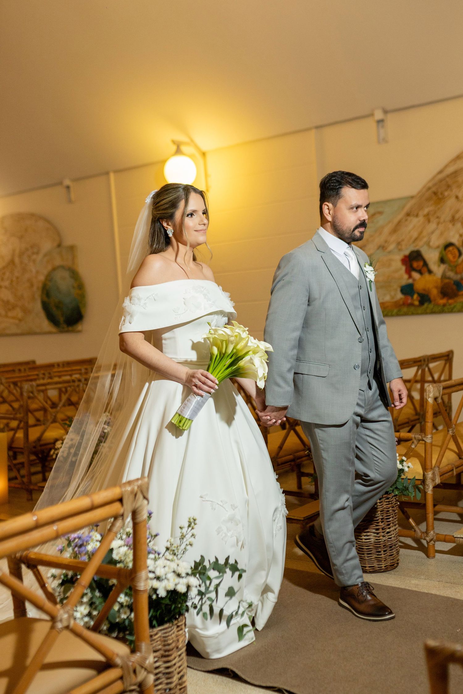 A bride in a white off-shoulder gown holds calla lilies while walking with a groom in a gray suit at an indoor wedding ceremony.