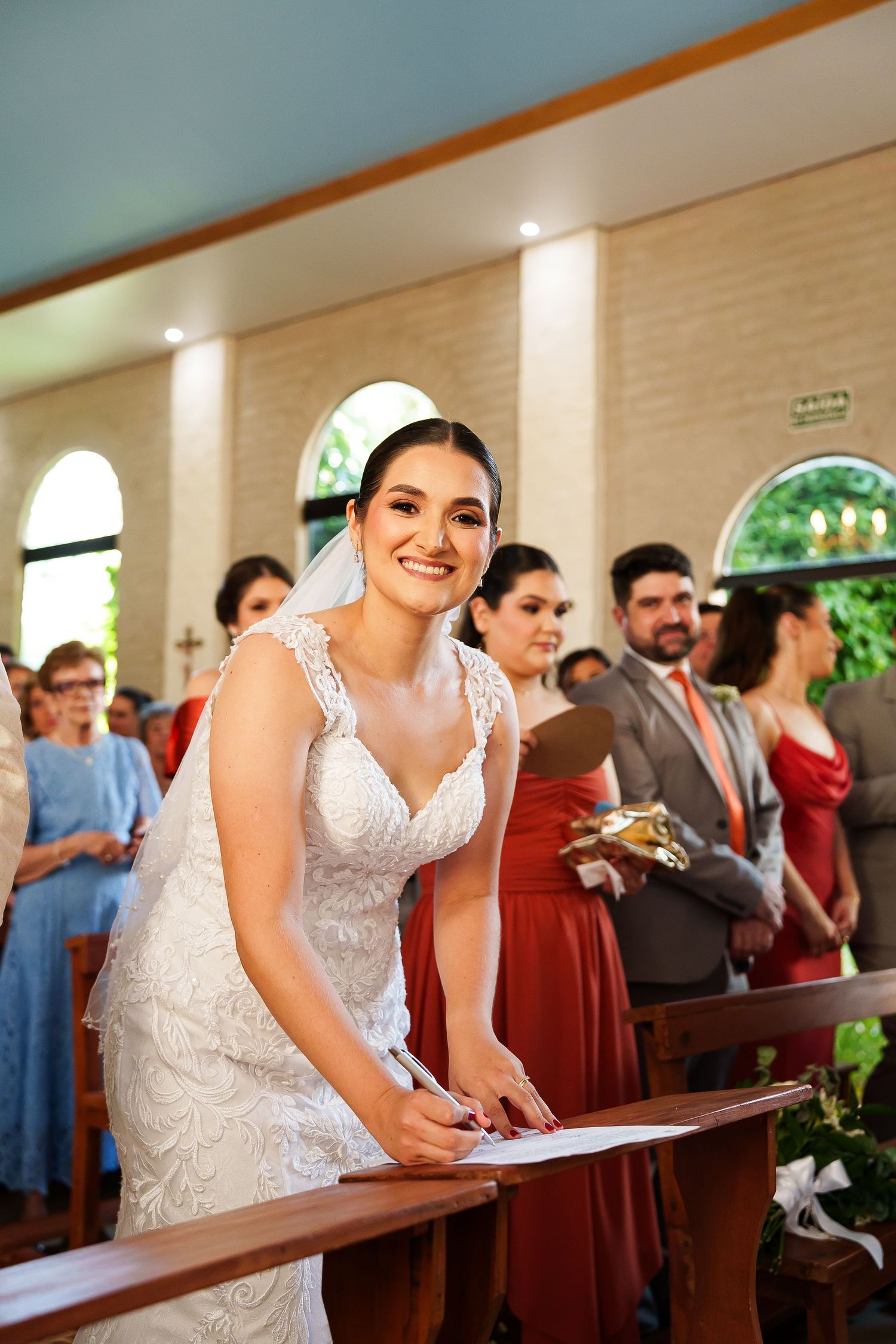 Smiling bride in white lace gown signing marriage certificate at church altar with wedding guests in background.