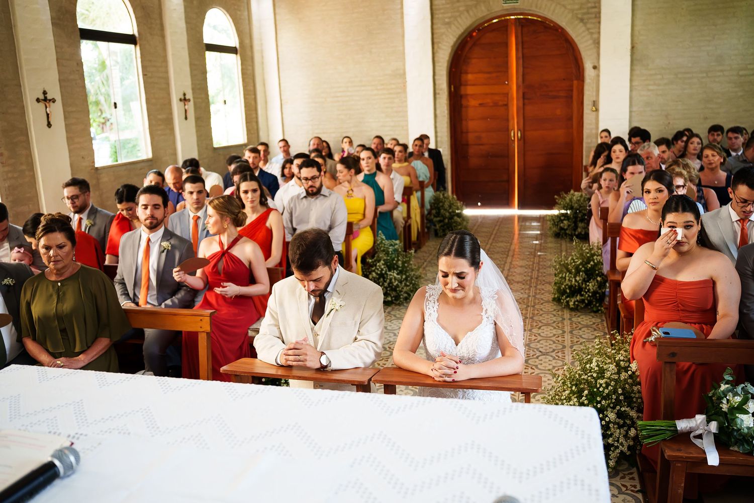 Bride and groom kneeling at church altar during wedding ceremony with guests seated in pews behind them.