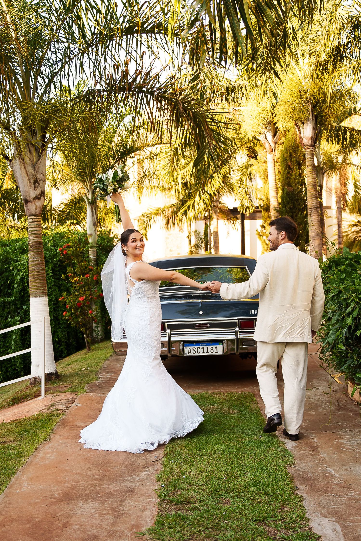 Bride in white gown and groom in beige suit hold hands near vintage car surrounded by palm trees on wedding day.
